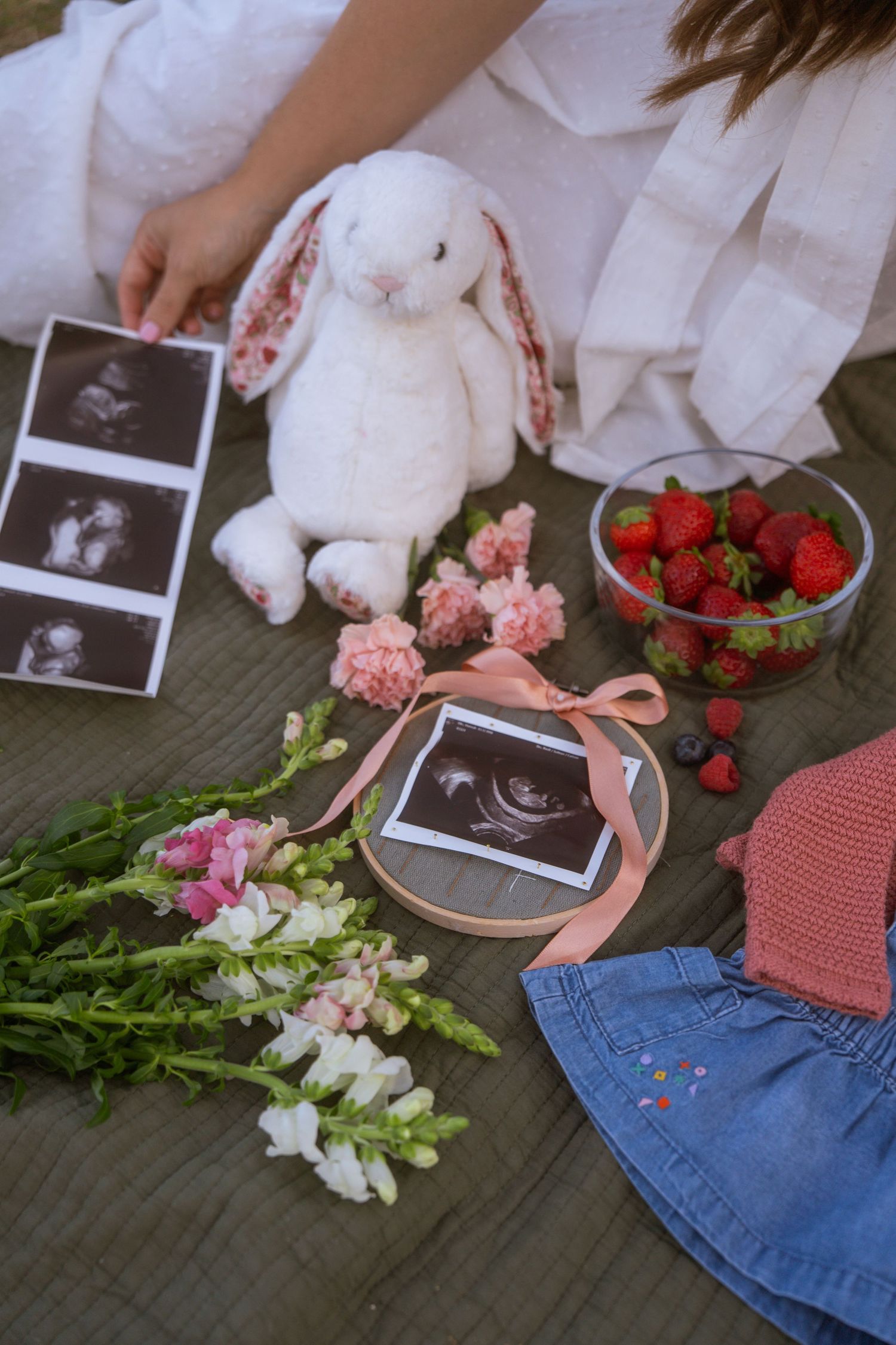 Maternity photo setup with ultrasound images, a white plush bunny, pink flowers, fresh strawberries and baby clothes on an olive blanket.