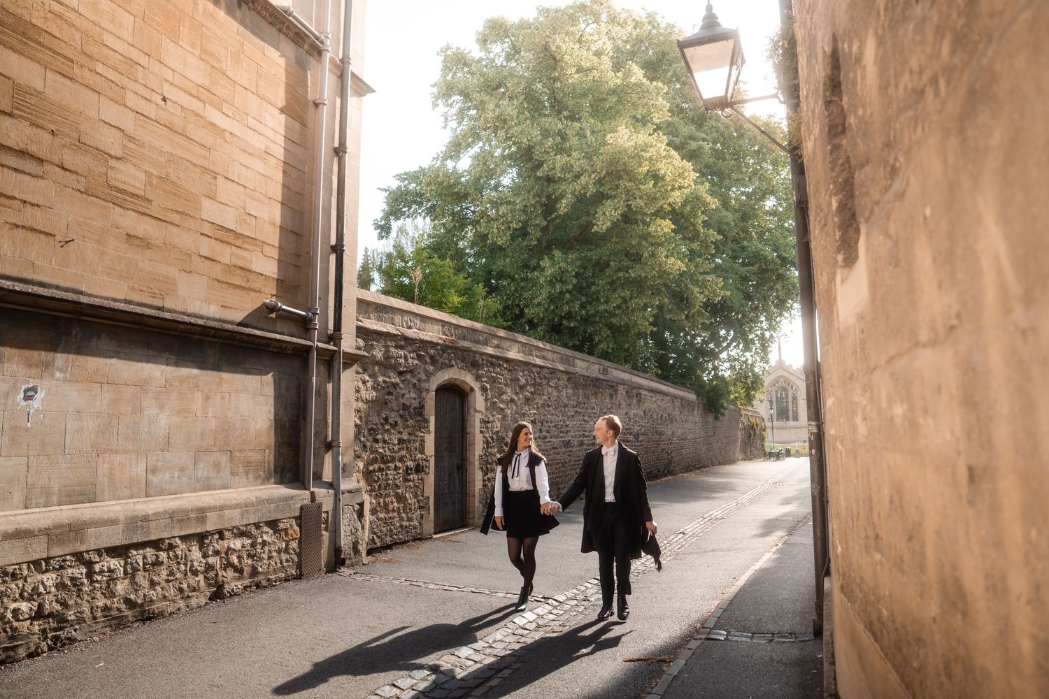Two people walk hand in hand down a sunlit cobblestone lane lined with historic stone walls and a leafy green tree.
