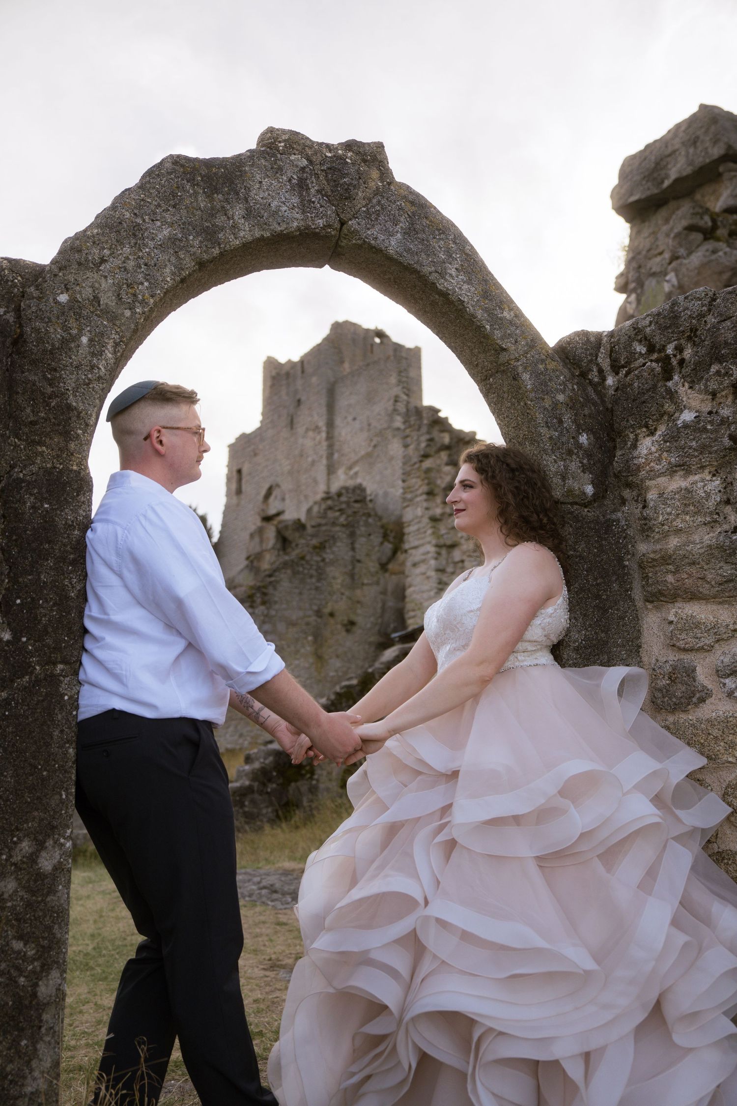A bride in a ruffled blush gown and a groom hold hands beneath a stone arch with castle ruins in the background.