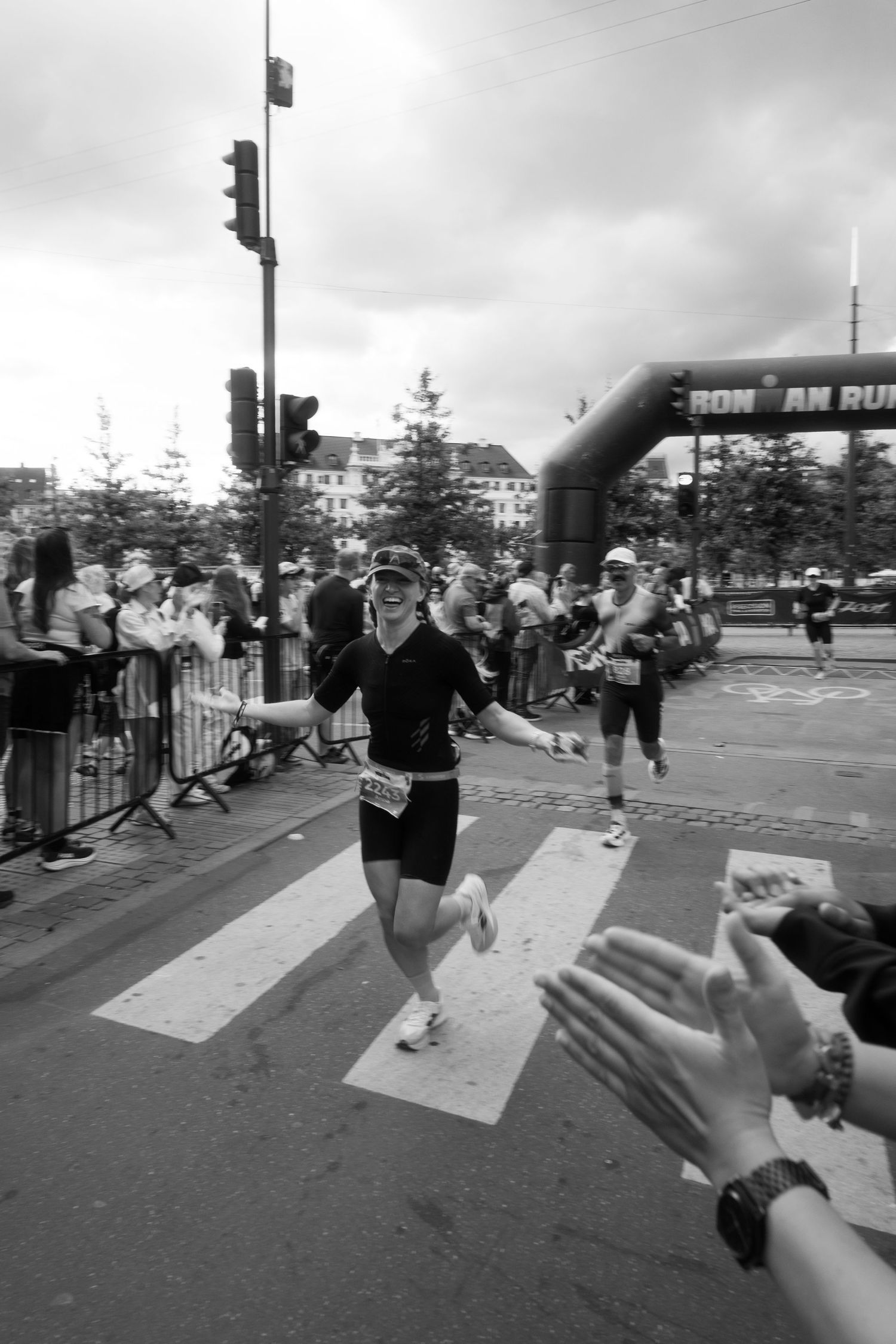 Smiling triathlete in black gear runs across a crosswalk past an Ironman arch, high-fiving cheering spectators in a black and white photo.