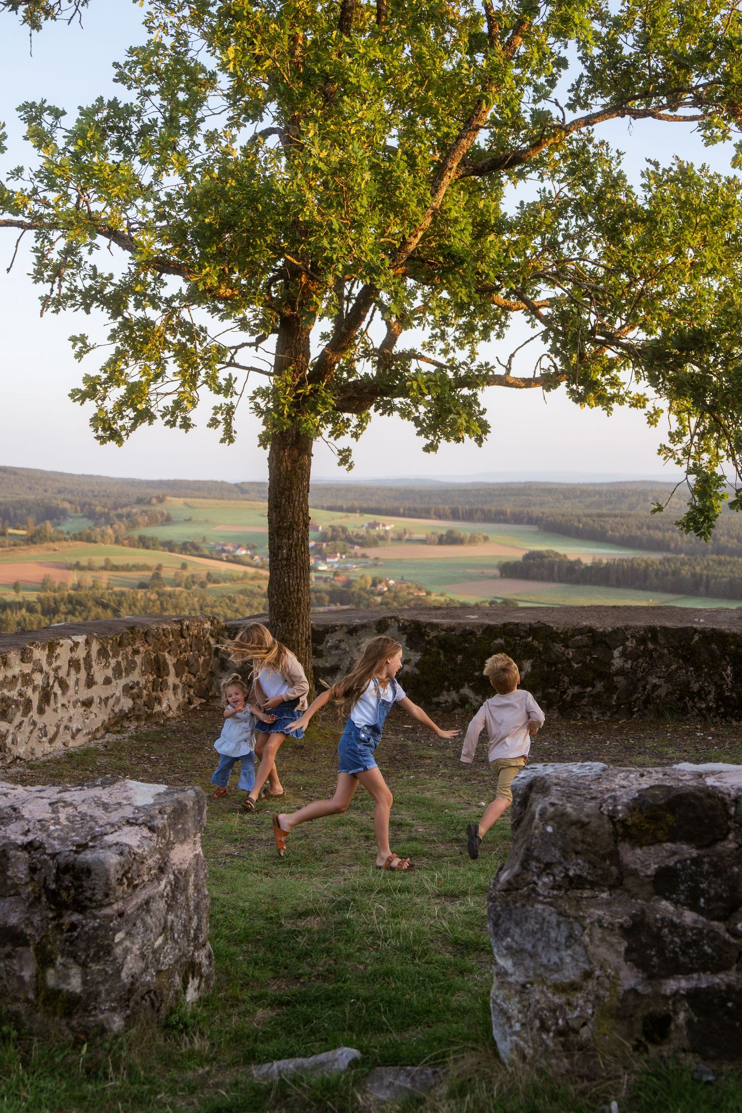 Children running and playing on a grassy hilltop beside old stone walls and an oak tree, overlooking rolling farmland.