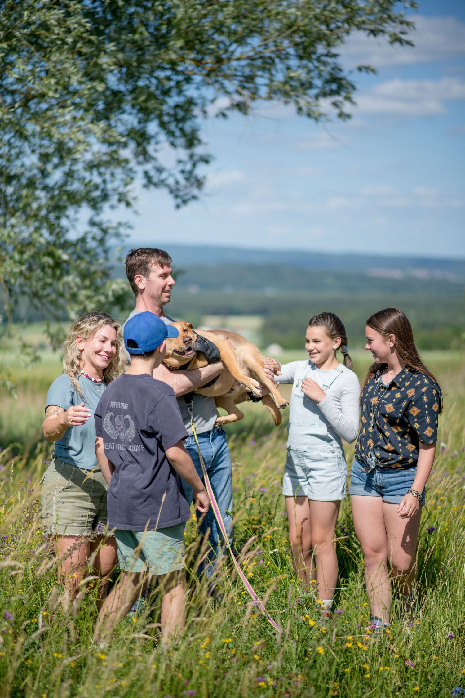 A smiling family of five stands in a sunny wildflower meadow while a man holds up a happy tan dog, with rolling hills behind them.