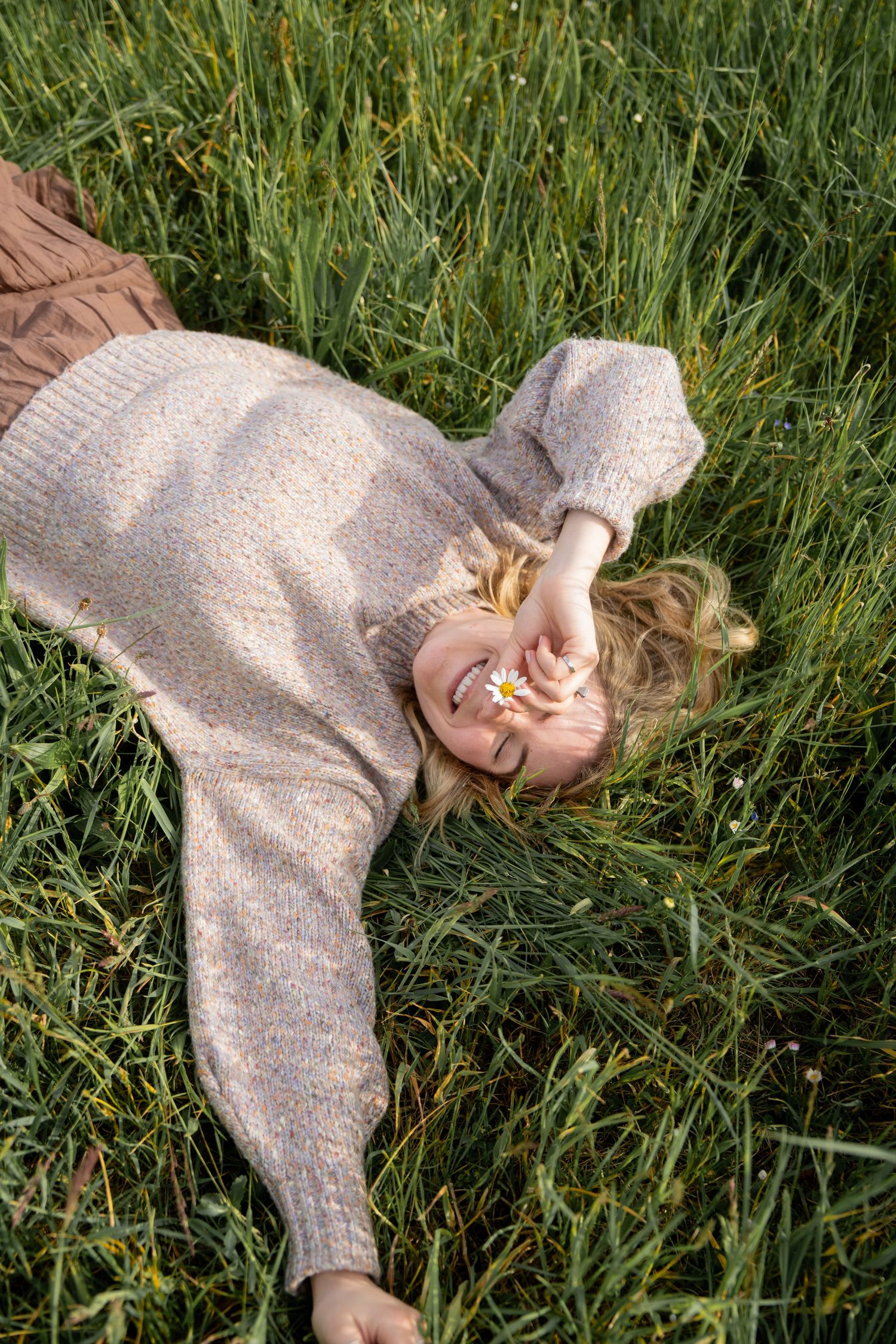 Smiling woman in a cozy knit sweater lies in tall green grass, holding a small white daisy over her eye on a sunny day.