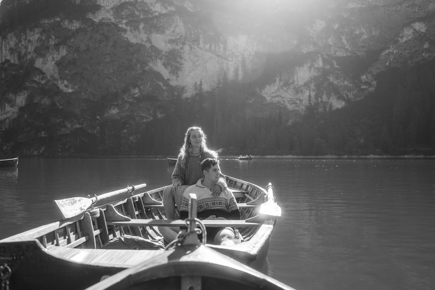 Two people sit in a wooden rowboat on calm mountain lake water as sunlight streams over steep rocky cliffs in a black and white photo.