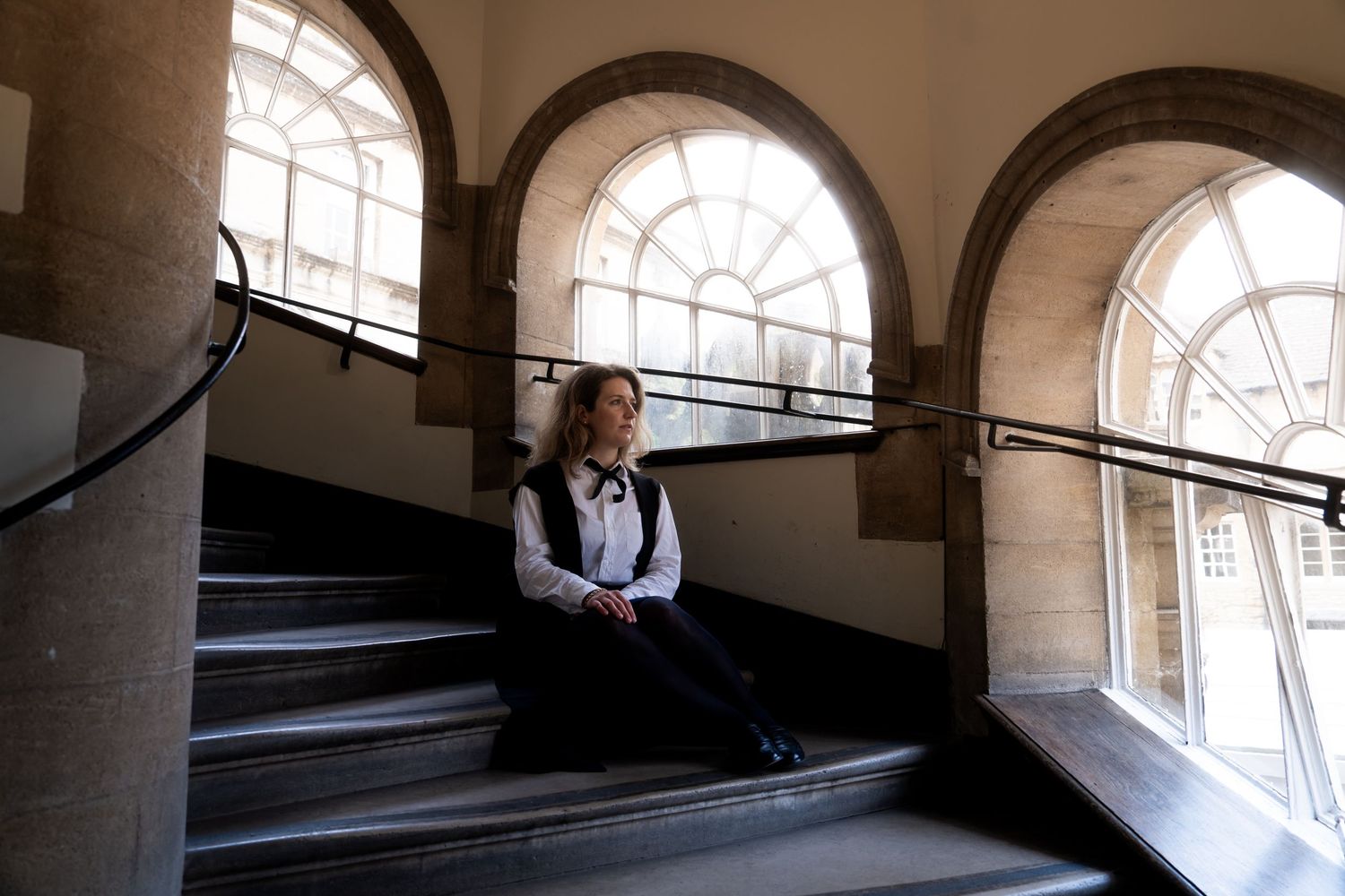 Woman in a white shirt and dark vest sits on a curved stone staircase beside tall arched windows flooded with natural light.