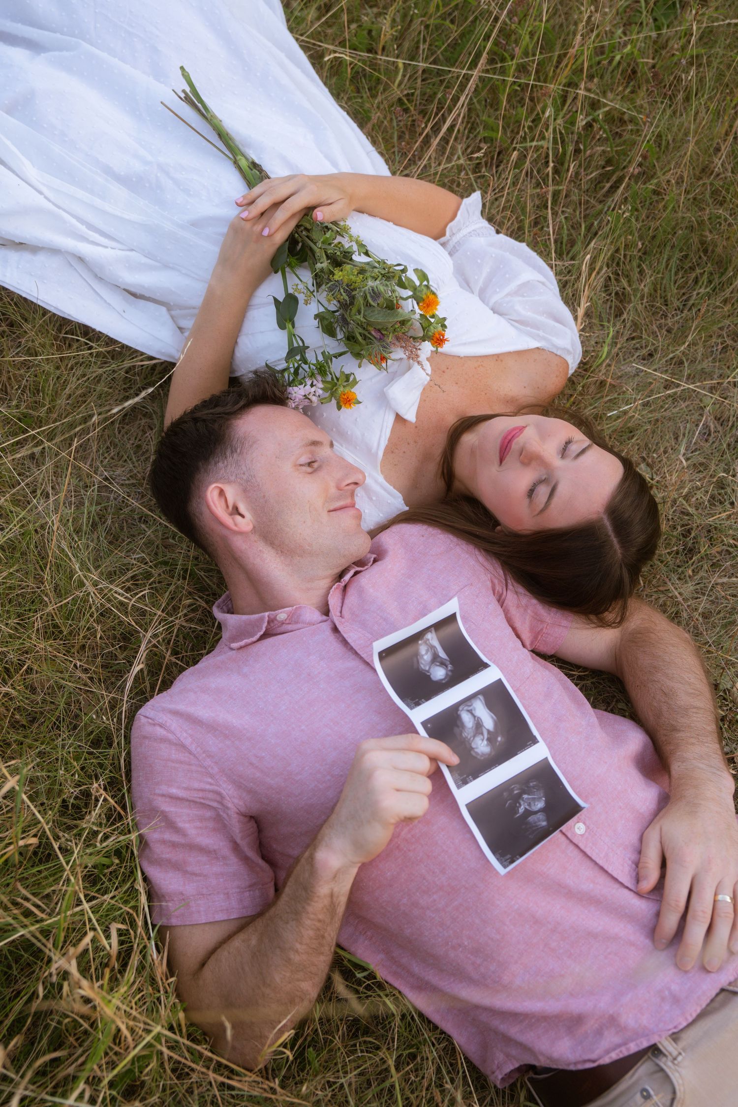 Expecting couple lying in a grassy field, holding ultrasound photos and a wildflower bouquet in a sweet maternity announcement.