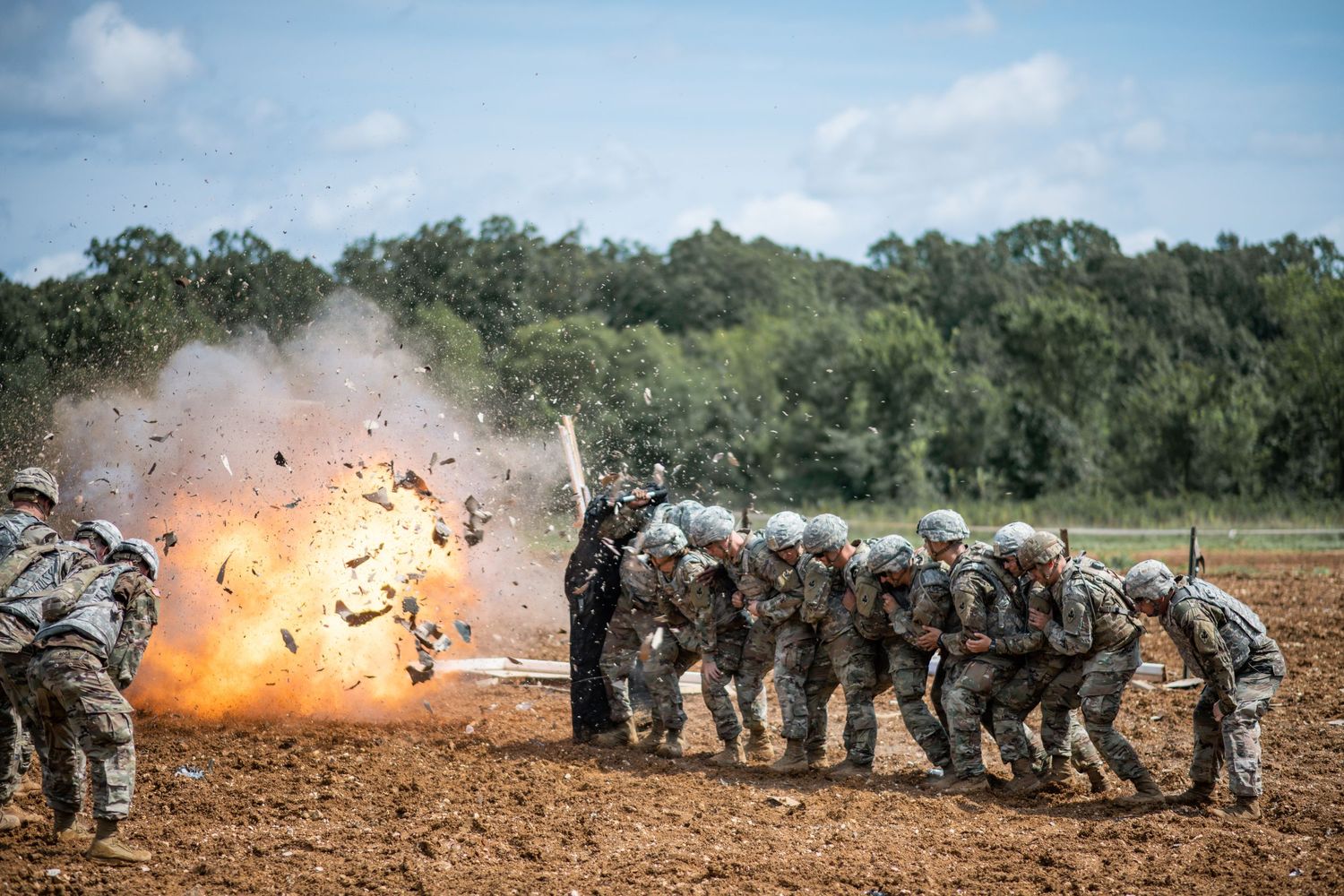Soldiers in camouflage uniforms and helmets huddle together beside a fiery explosion during outdoor military training.