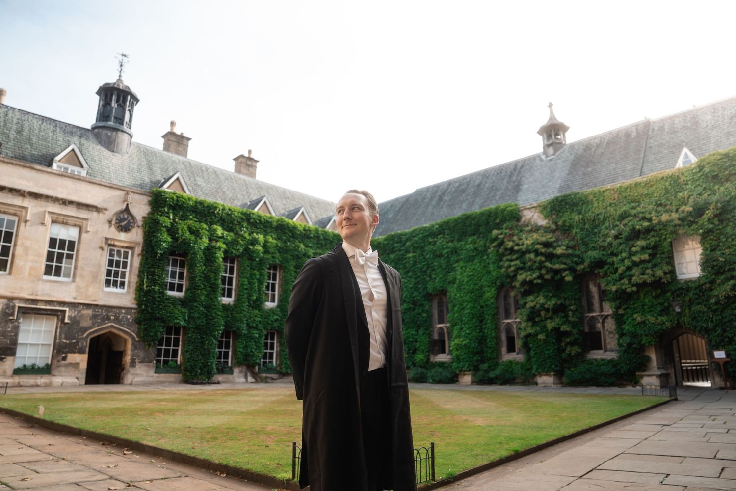 A person in a black academic gown stands in a sunlit college courtyard surrounded by ivy-covered stone buildings.