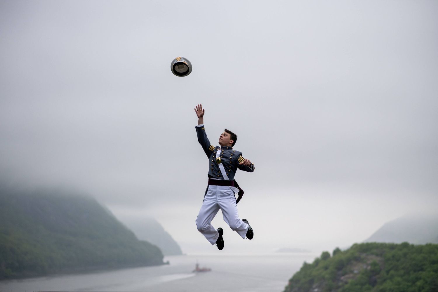 A uniformed cadet leaps high into the air tossing a cap above a misty river valley with fog-covered hills.