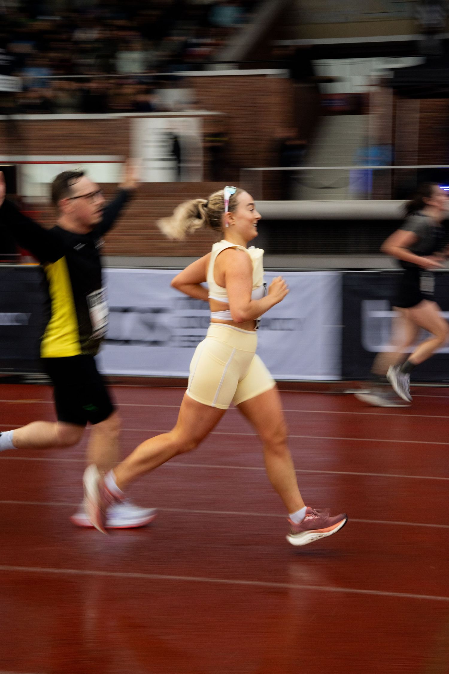 A blonde woman in a cream sports outfit sprints around an indoor red running track beside another runner in motion blur.