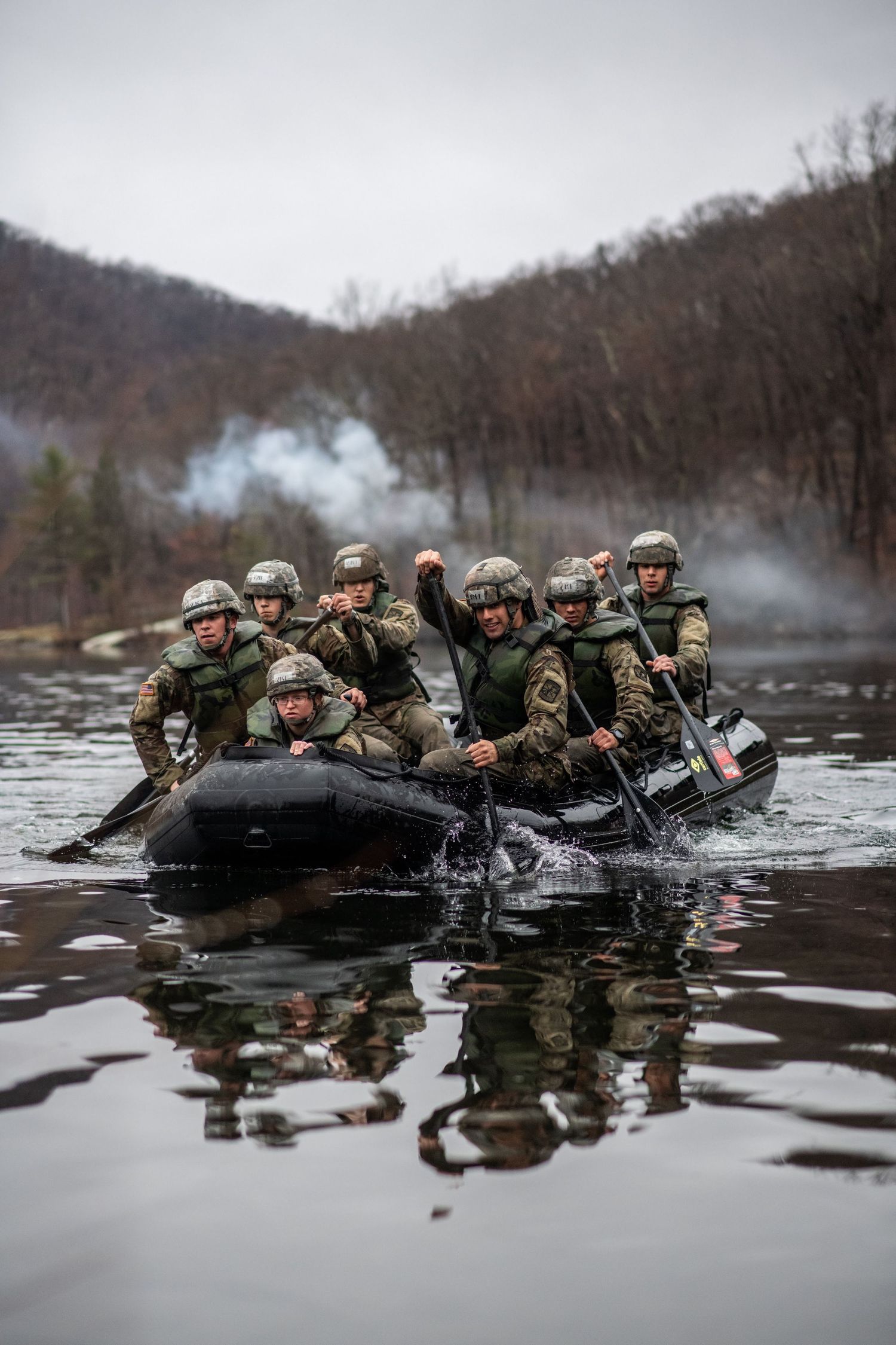 A team of helmeted soldiers paddles an inflatable raft across a calm lake with smoke rising from wooded hills.