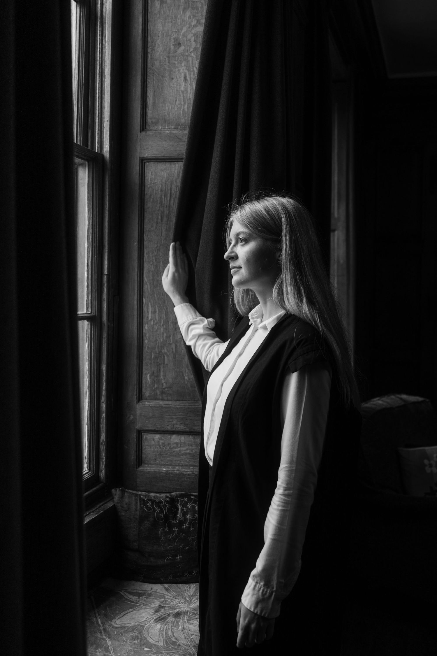 Black and white portrait of a woman with long hair in a dark vest and white blouse, holding a heavy curtain while gazing out a window.