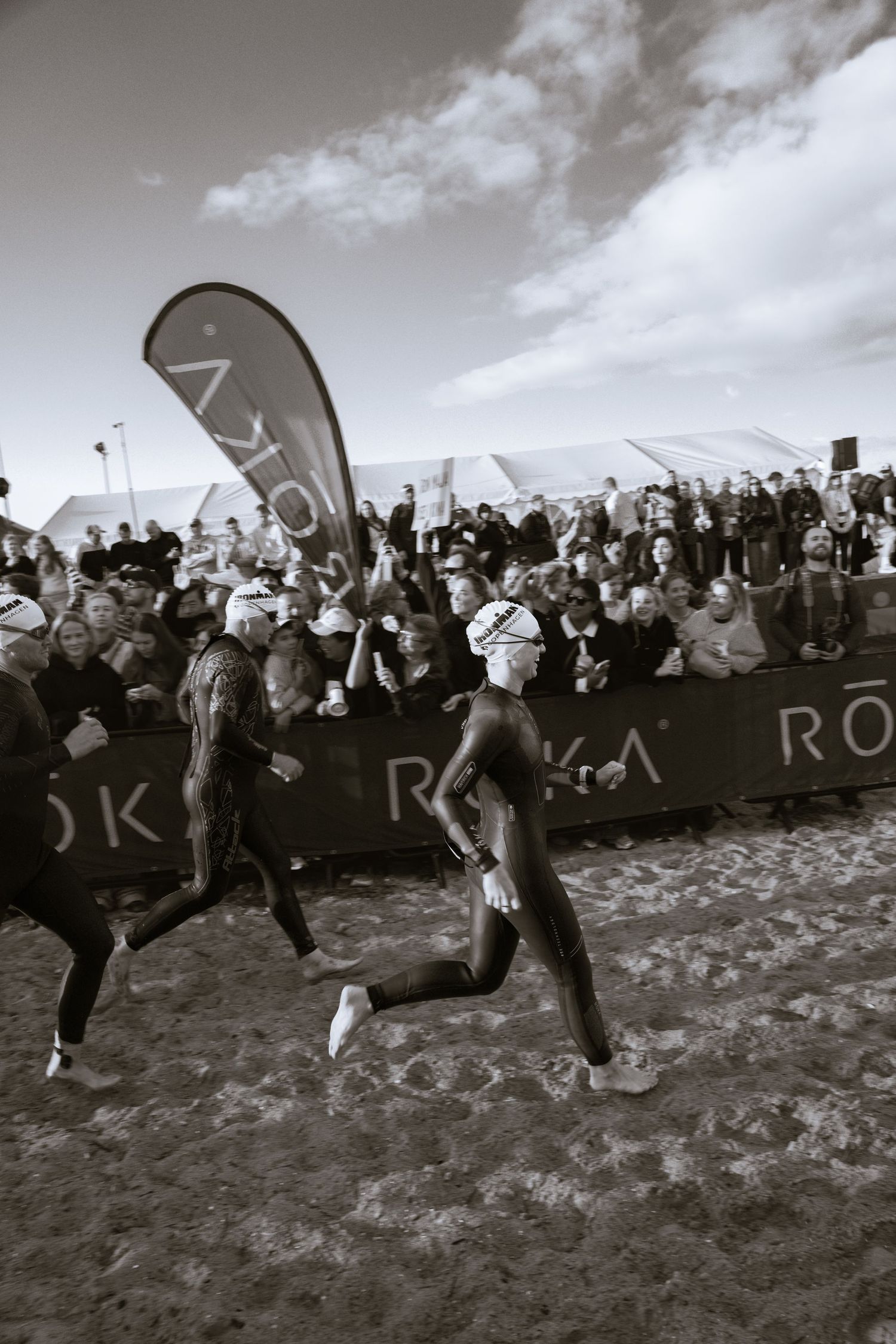 Triathletes in wetsuits run across the sand past cheering spectators and Roka sponsor banners in a black and white race scene.