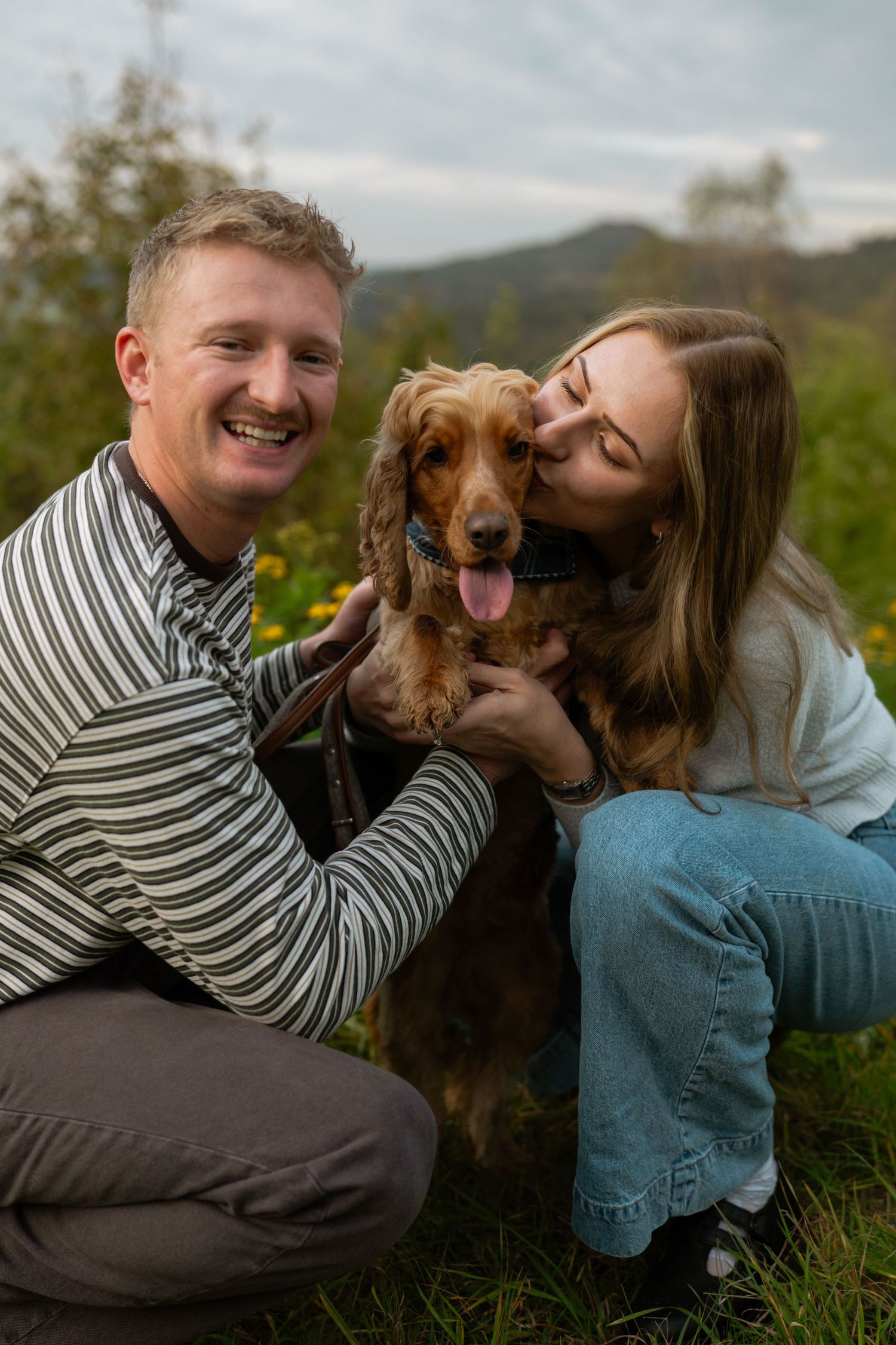 Smiling man and woman kneel in a grassy field as a golden cocker spaniel licks the woman's cheek.