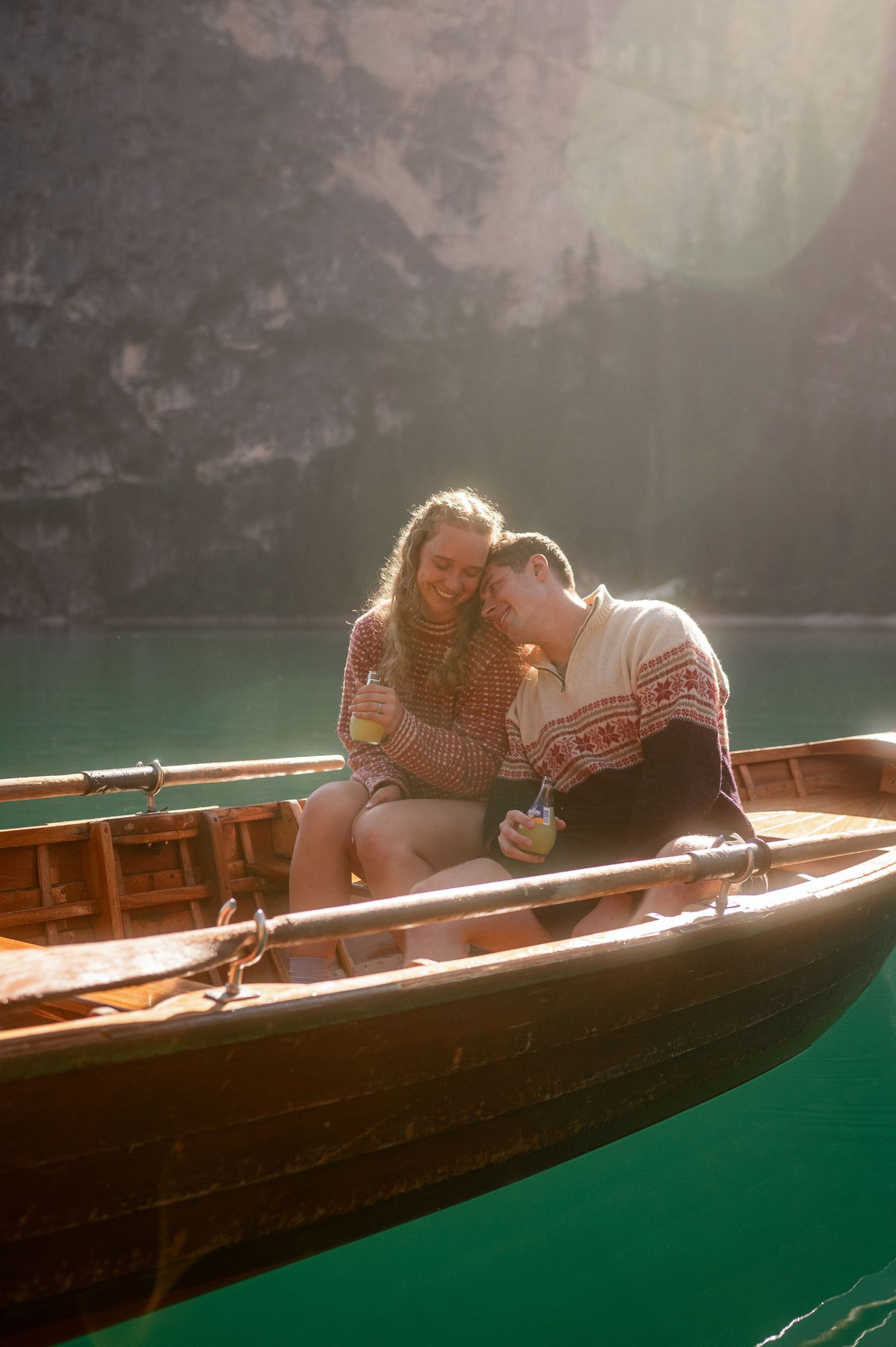 Couple in matching knit sweaters cuddle and toast with drinks in a wooden rowboat on calm emerald lake beneath sunlit mountain cliffs.