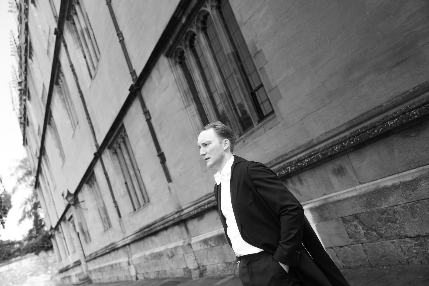 Black and white photo of a man in a dark suit and white shirt standing beside a historic stone building with tall arched windows.