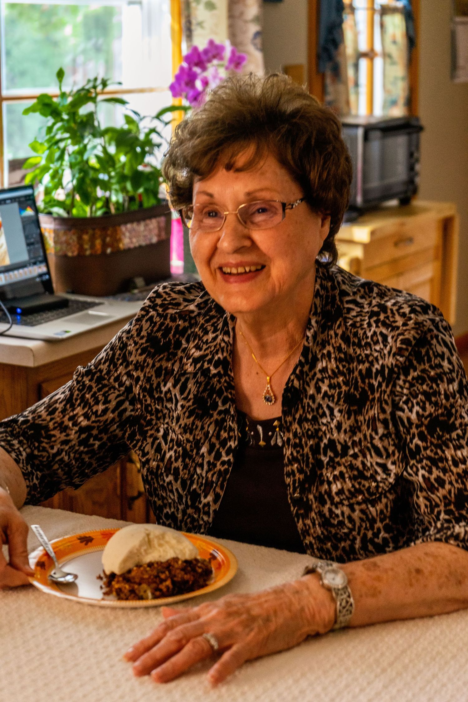 Smiling woman in animal print cardigan sits at dining table with plate of food and houseplants in background.
