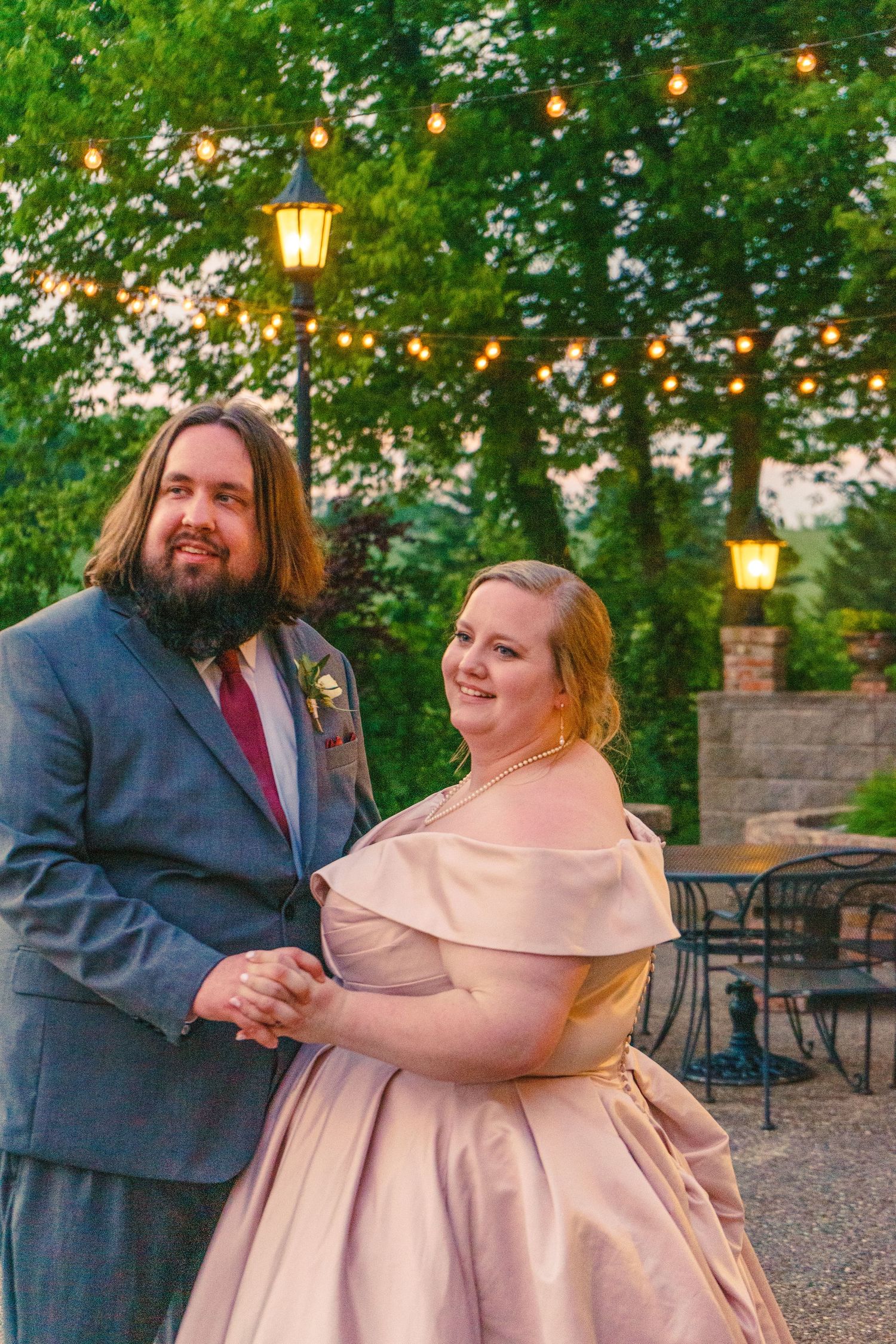 A couple stands outdoors at twilight under string lights and lanterns at a garden venue dressed in formal attire.