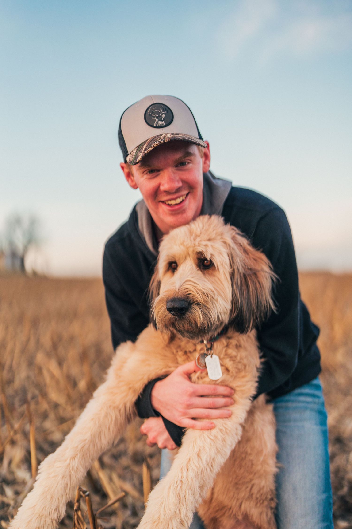 A shaggy golden Goldendoodle dog poses in a brown field with someone wearing jeans and a dark jacket on a sunny day.