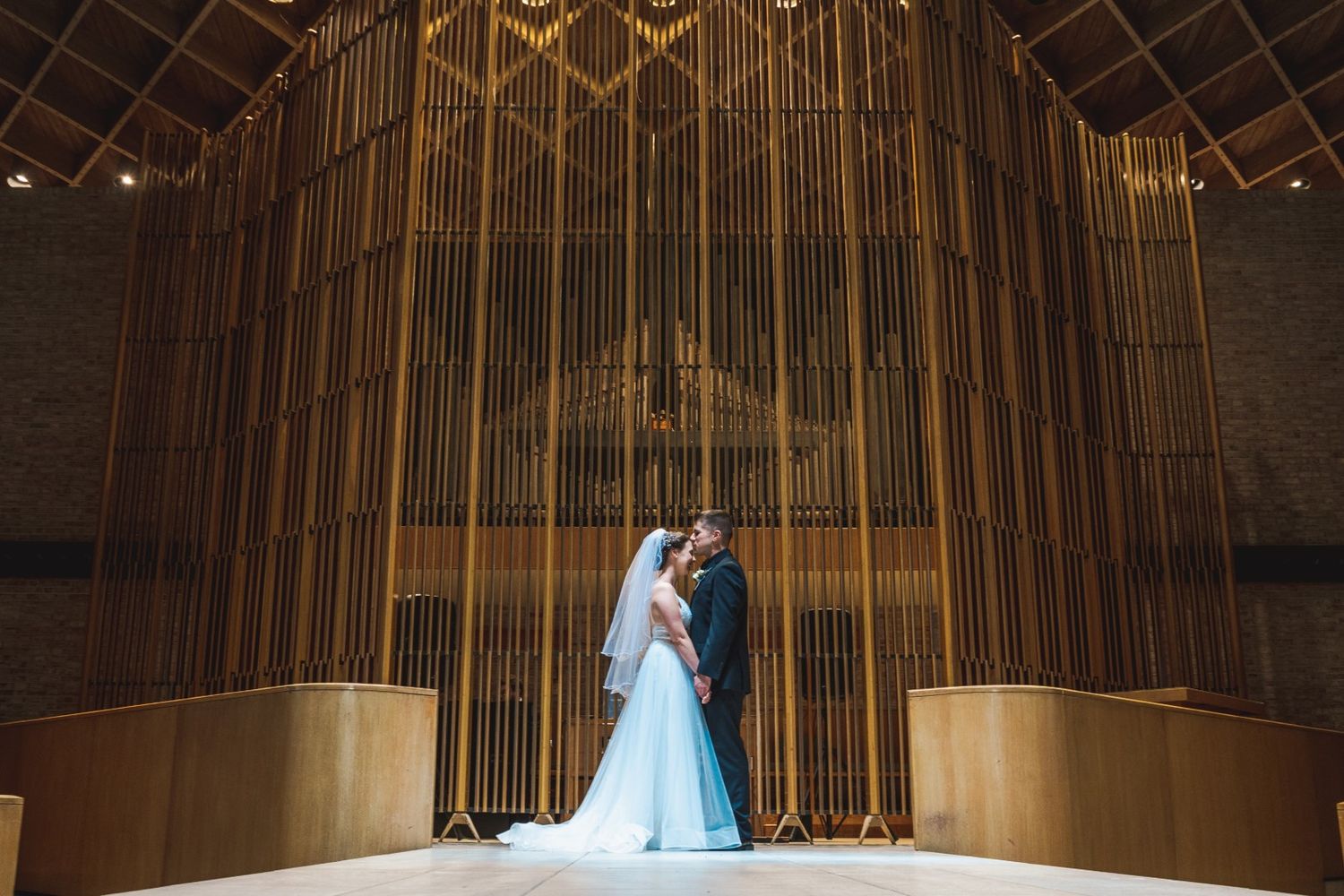 A wedding couple stands in dramatic lighting against ornate golden organ pipes in a grand hall.