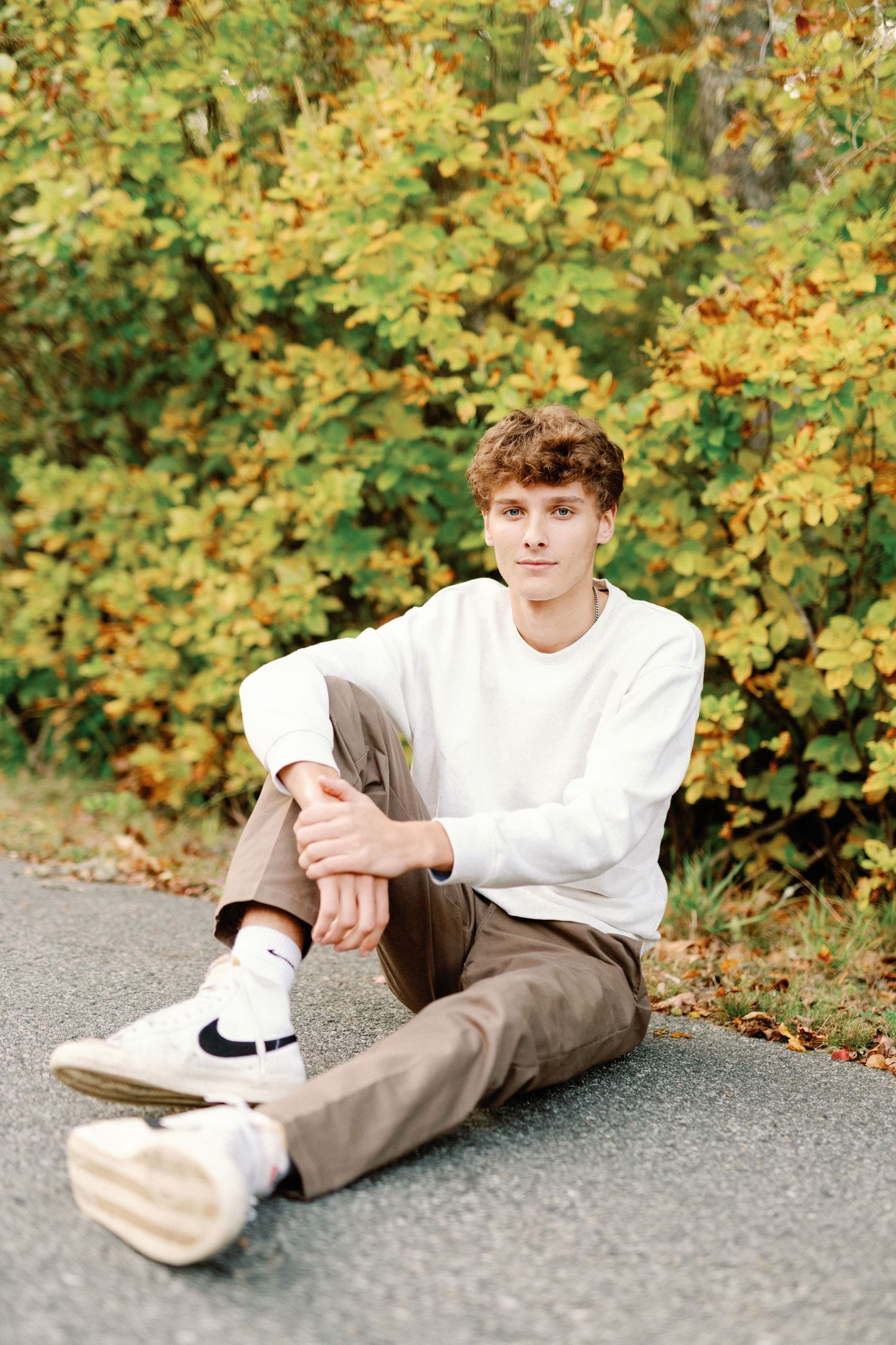 A person in a white sweater and khaki pants sits casually on pavement with autumn foliage in the background.