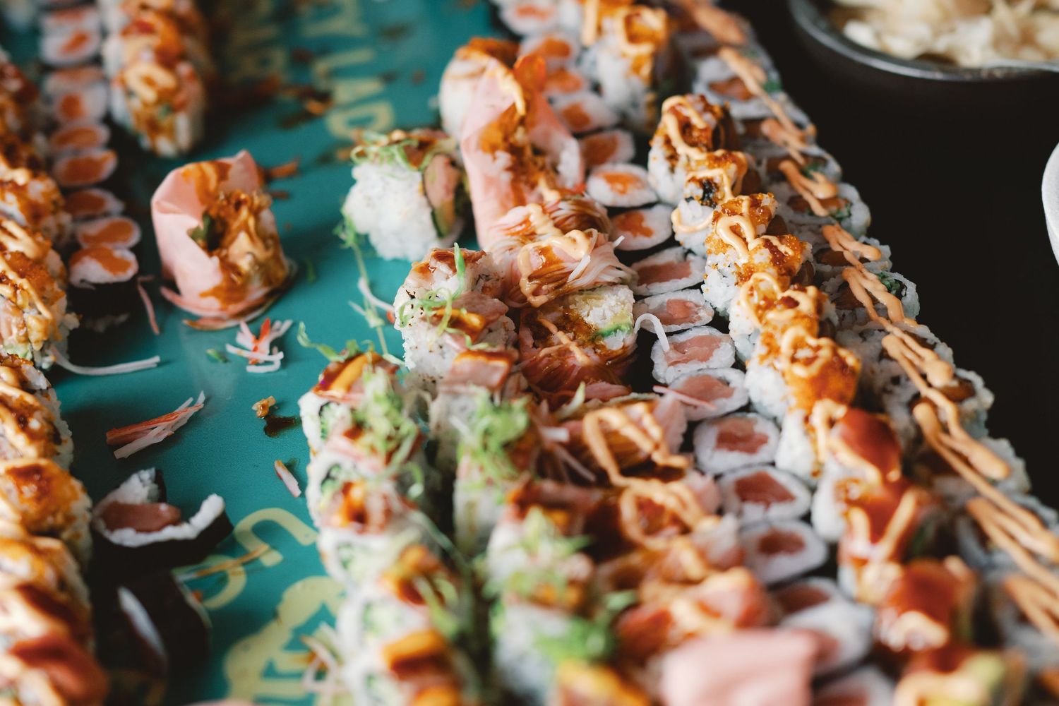 Rows of sushi rolls topped with spicy sauce and garnished with green herbs arranged on a dark surface.