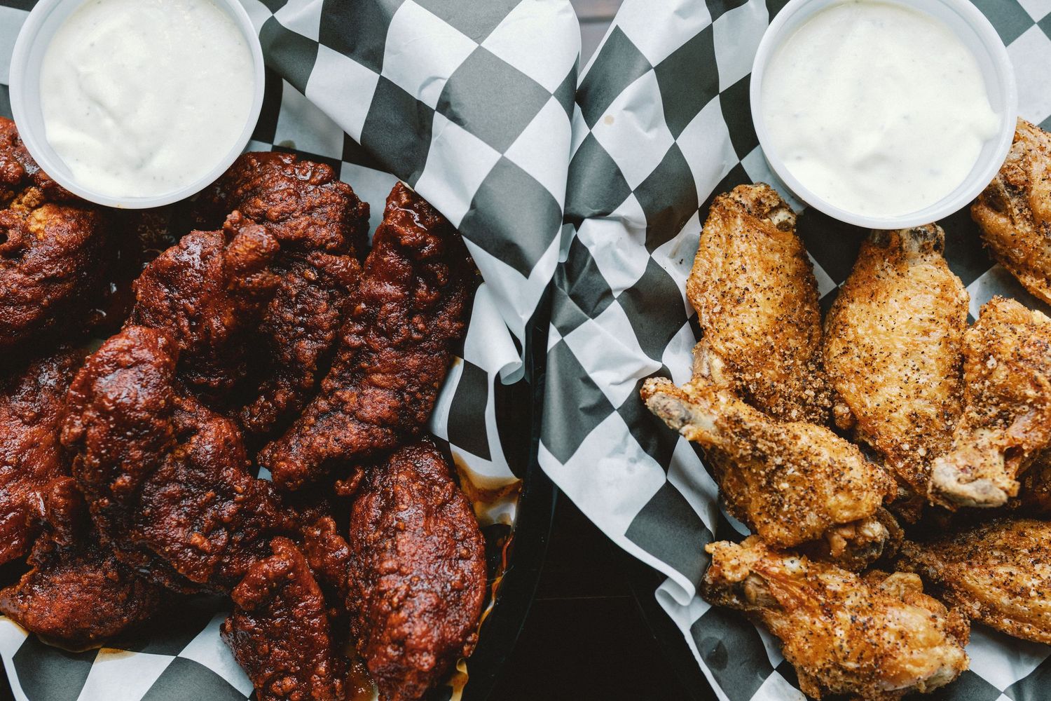 Assorted chicken wings with different sauces and dips served in black and white checkered paper baskets.