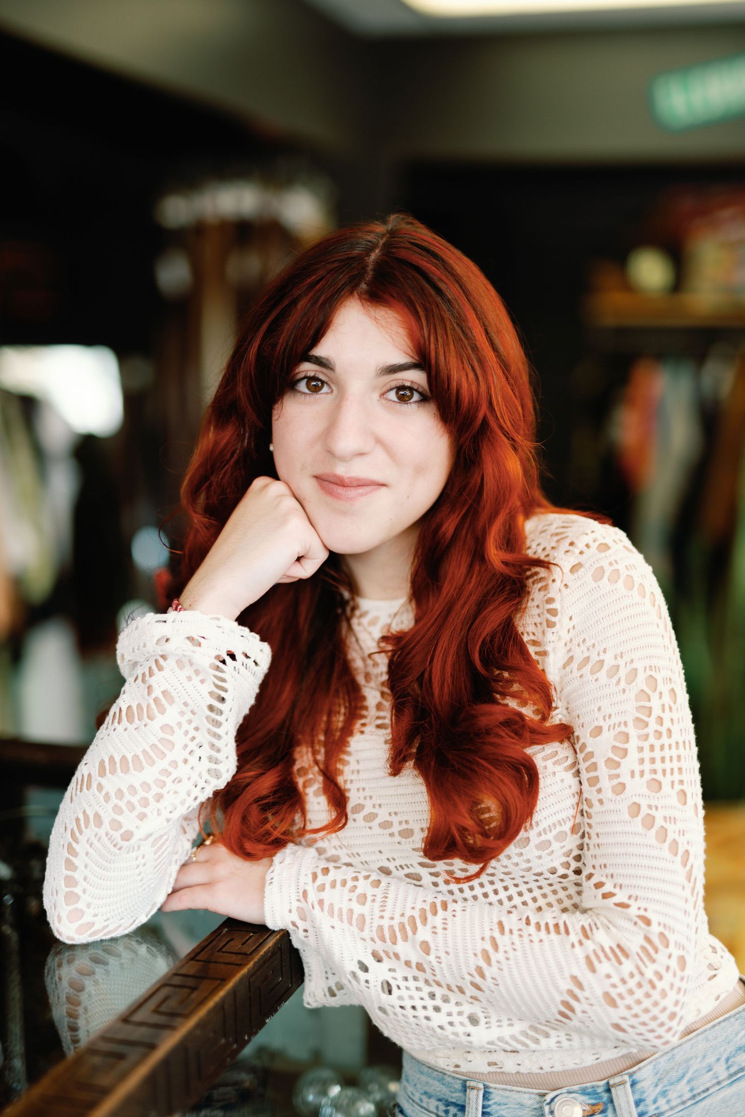 A person with red hair wearing a white crochet top poses in a casual indoor setting.