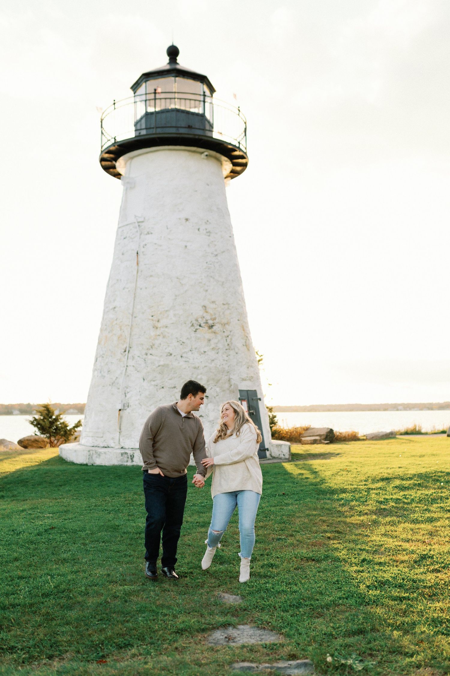 A couple stands together on green grass in front of a tall white lighthouse on a coastal landscape.