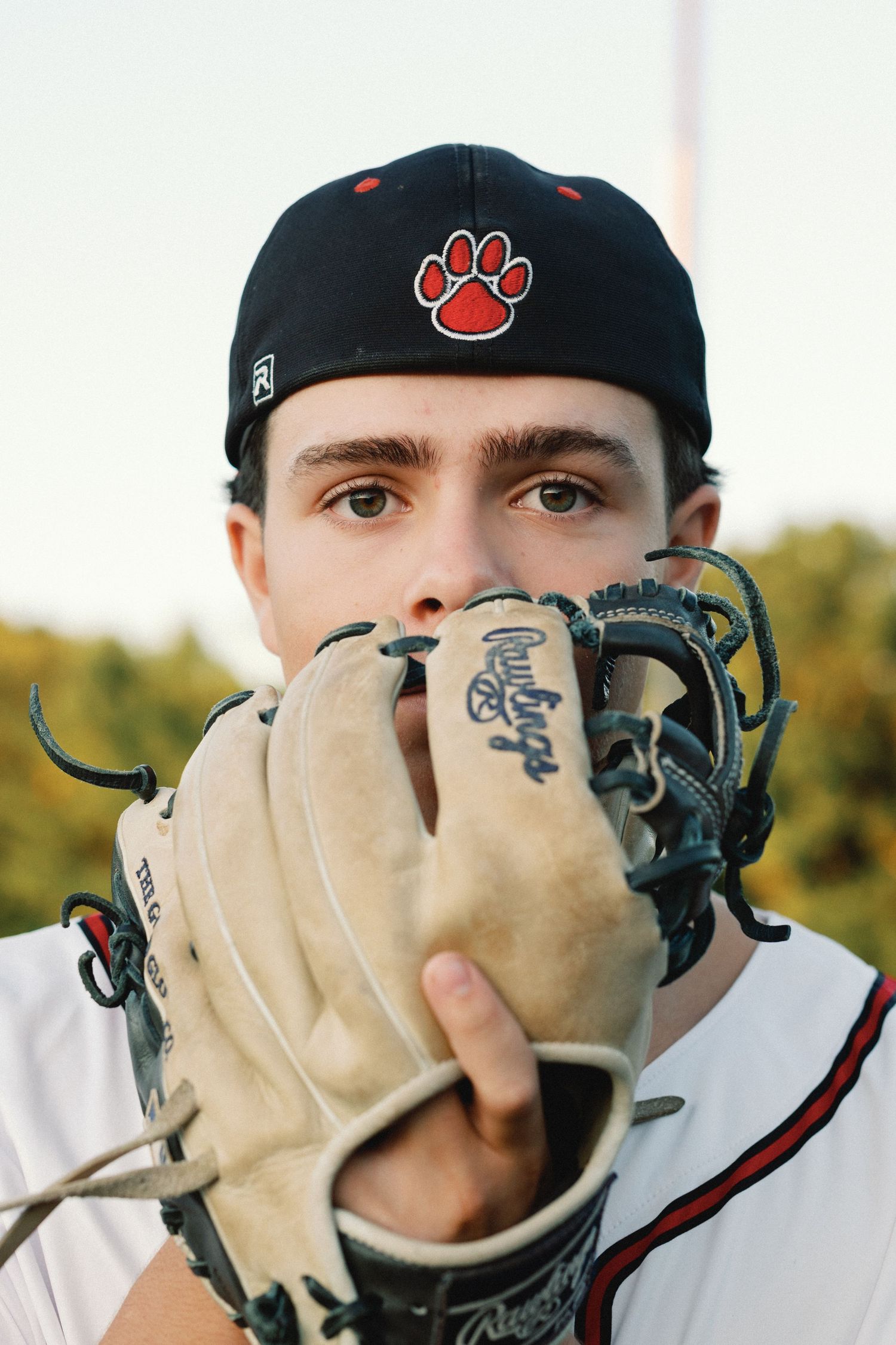 Baseball player wearing black cap with red paw logo holds up leather glove in front of face during outdoor photo.