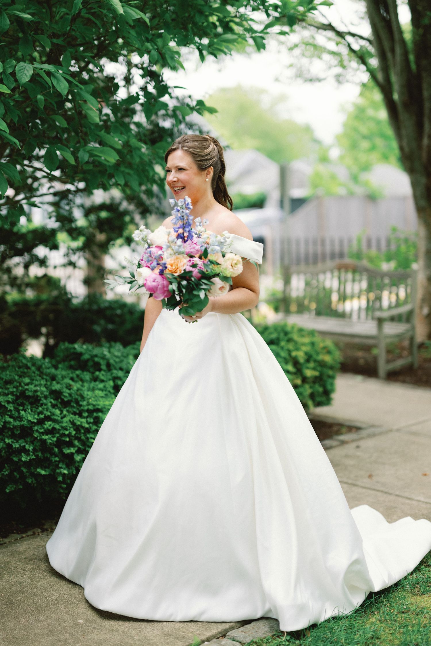 A beautiful off-shoulder white wedding gown with a full skirt and colorful floral bouquet in a garden setting.