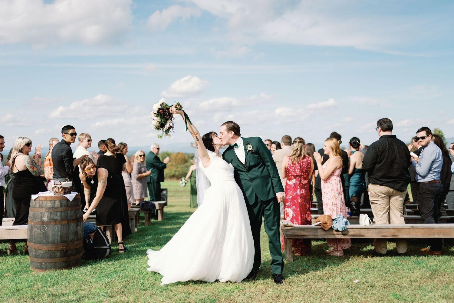 A couple celebrates during an outdoor wedding ceremony with guests gathered around wine barrels on a grassy hilltop.