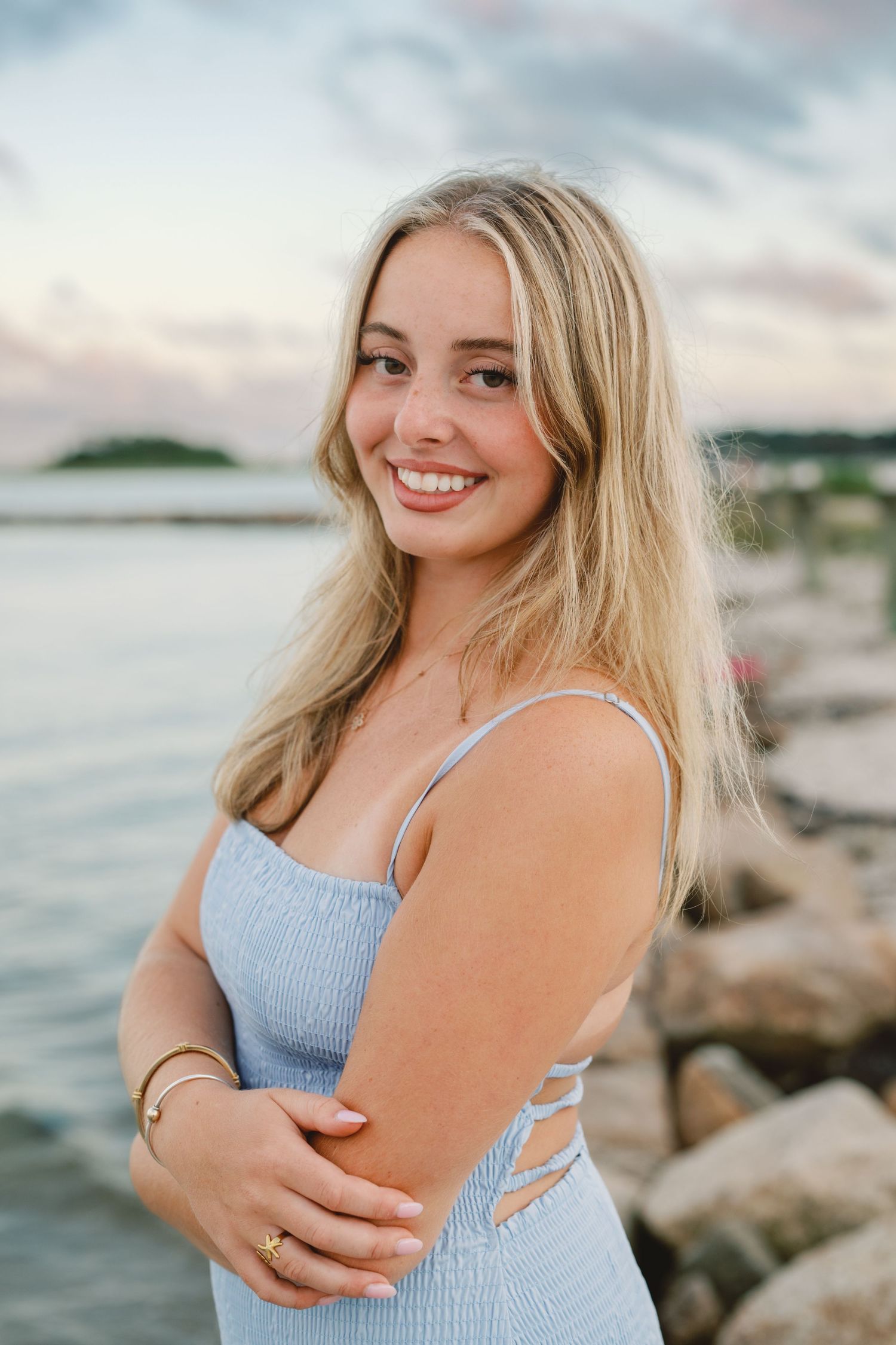 Smiling blonde woman in a light blue sundress posing by rocky shoreline at sunset.