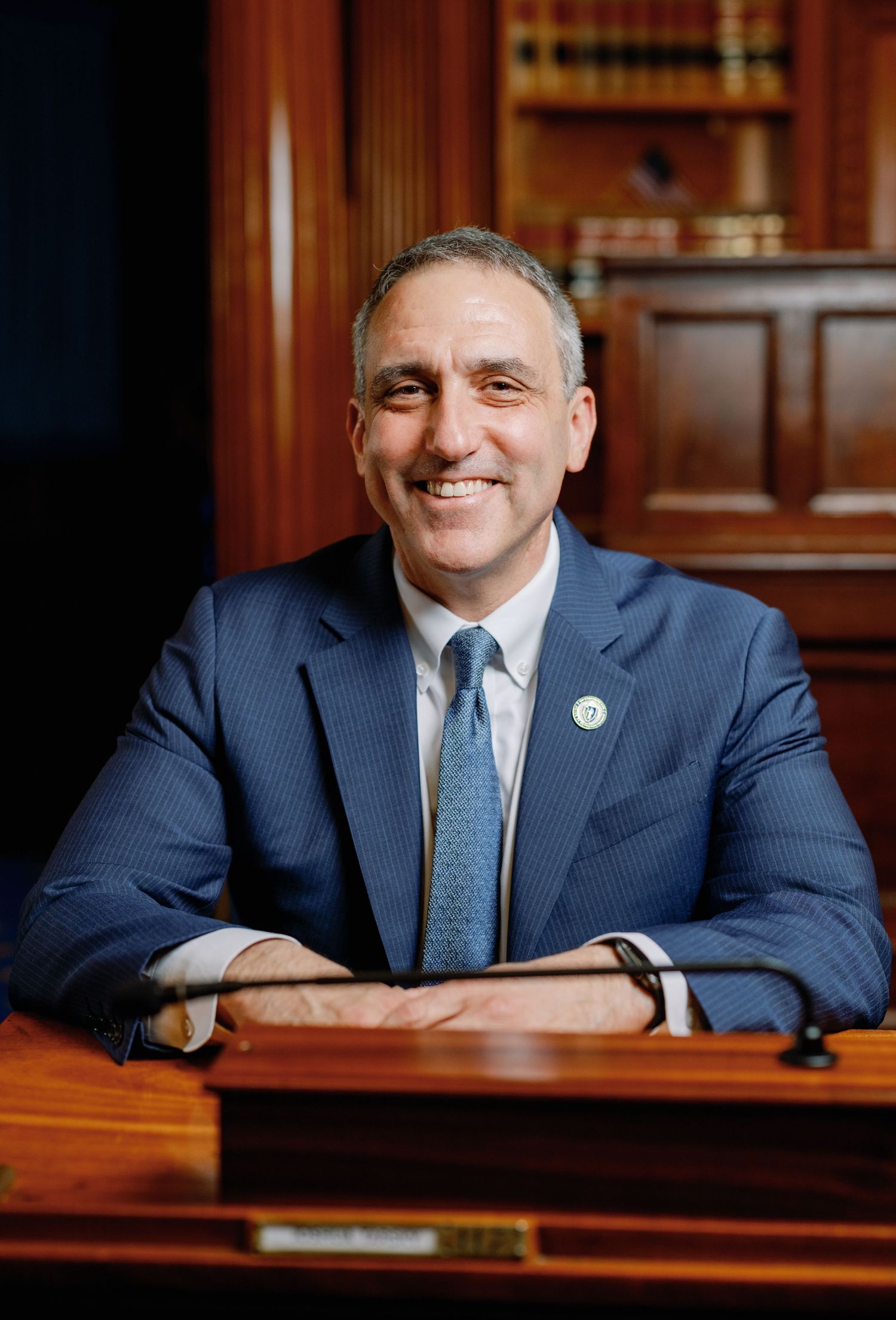 Professional man in blue suit smiling at wooden desk with law books in background, formal office portrait.