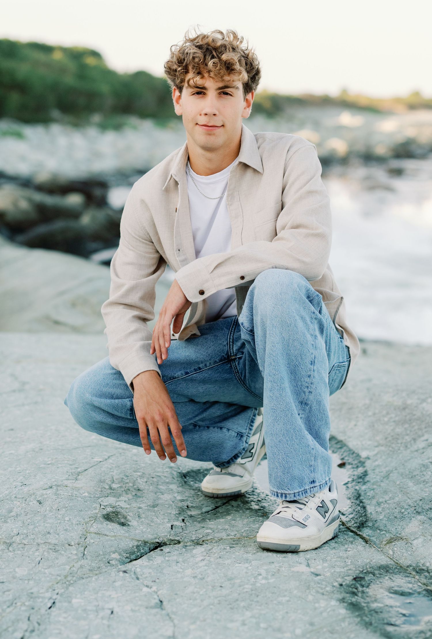 Casual portrait of someone in beige jacket and jeans sitting on rocky beach at sunset.