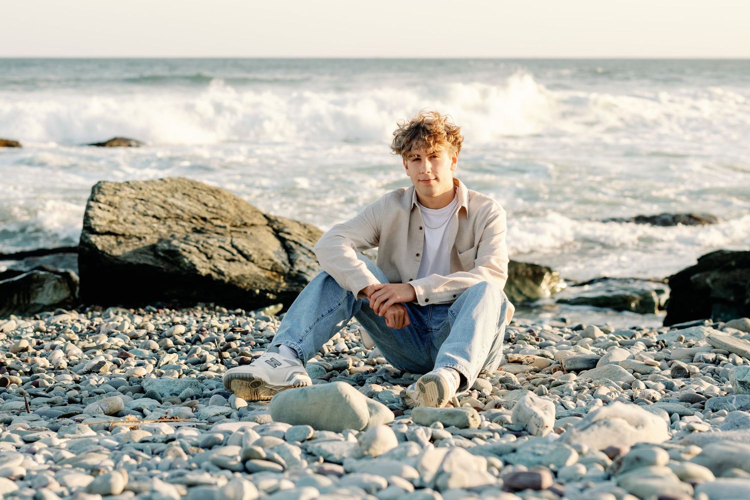 Young man with curly hair sitting on rocky beach, wearing casual outfit, with ocean waves and boulders in background.