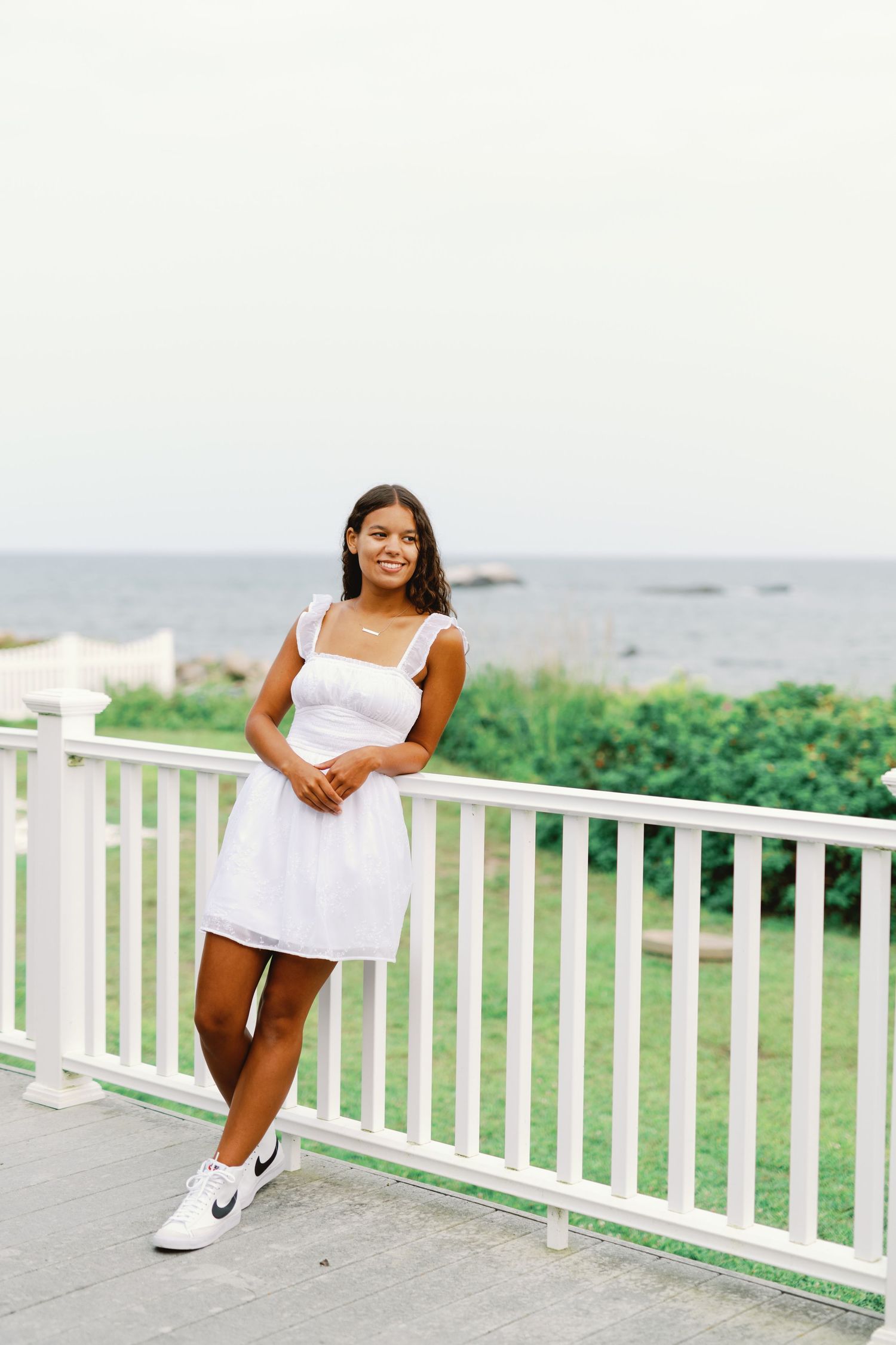 Person in a white sundress and sneakers poses on a porch overlooking the ocean and greenery.