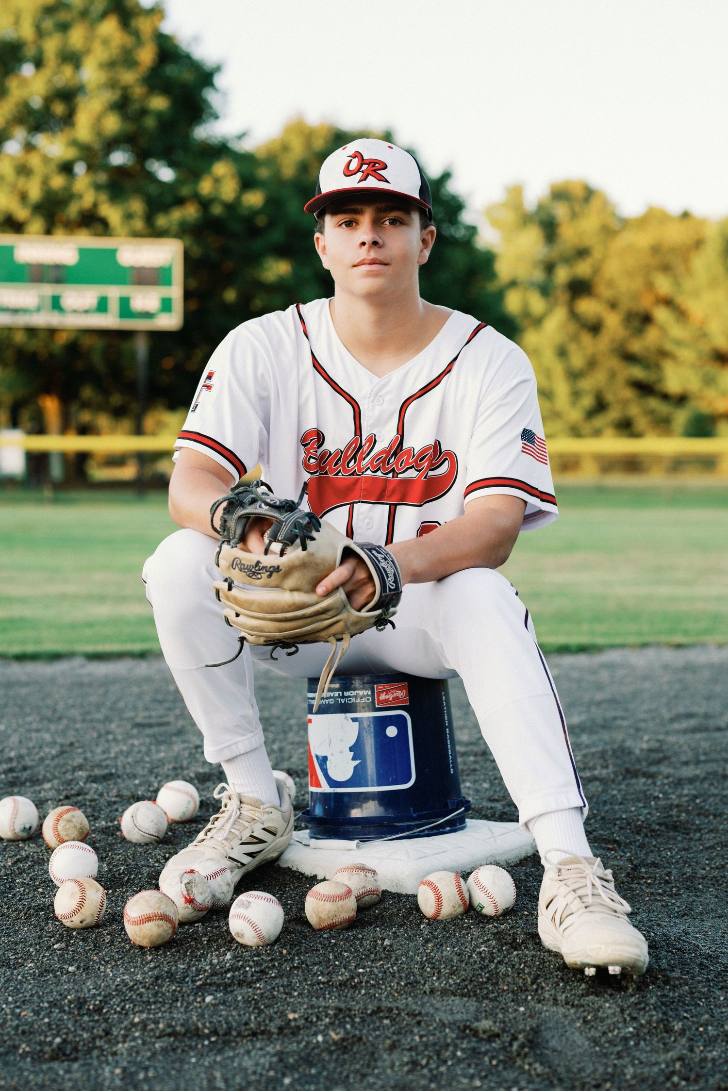 Baseball player in white uniform sits with glove and baseballs on field during golden hour photoshoot.