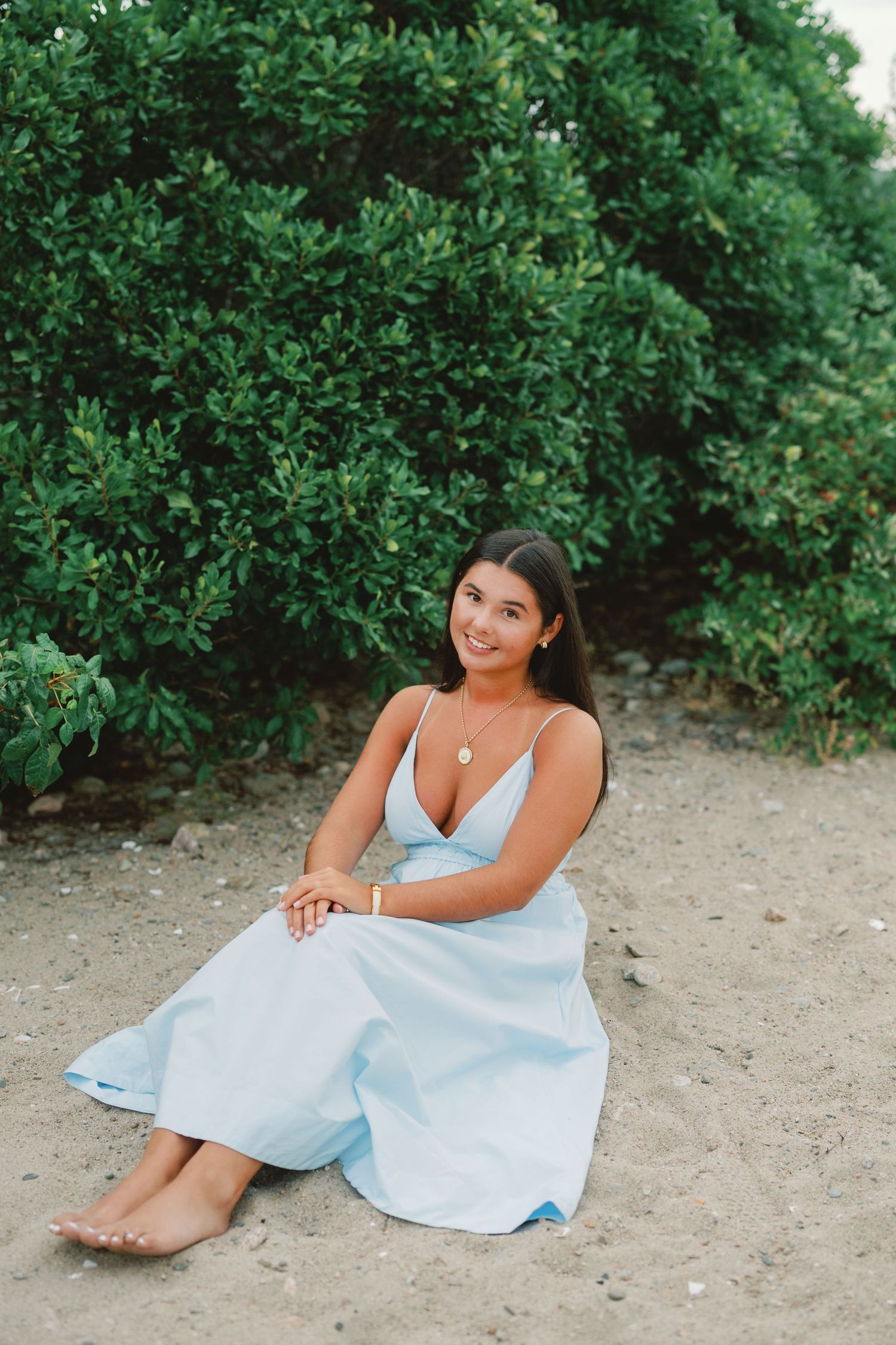 Young woman in light blue dress sitting on sandy ground in front of lush green bushes, smiling gracefully.
