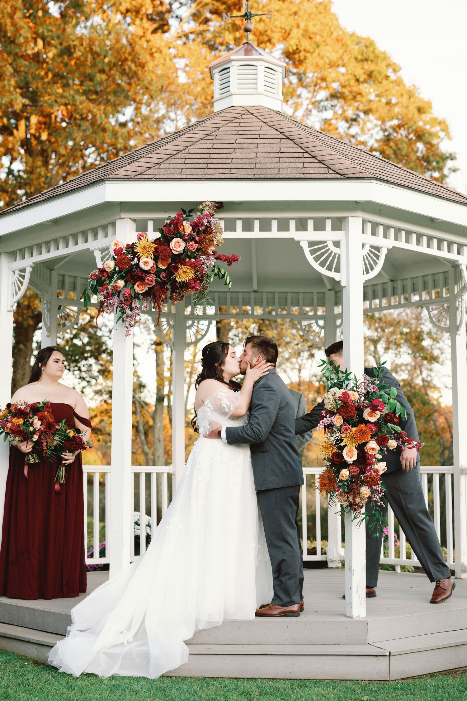 Fall wedding ceremony takes place in a white gazebo decorated with burgundy and pink floral arrangements.