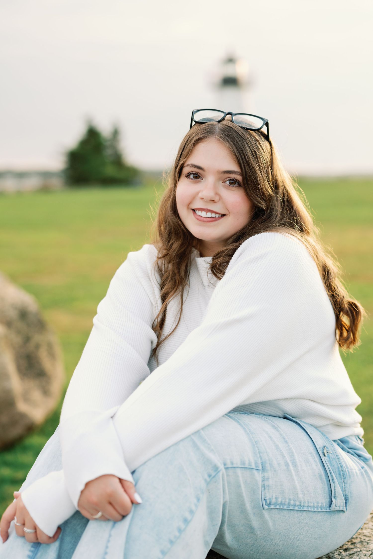 A person in a white sweater and light blue jeans sits casually outdoors in a grassy field.