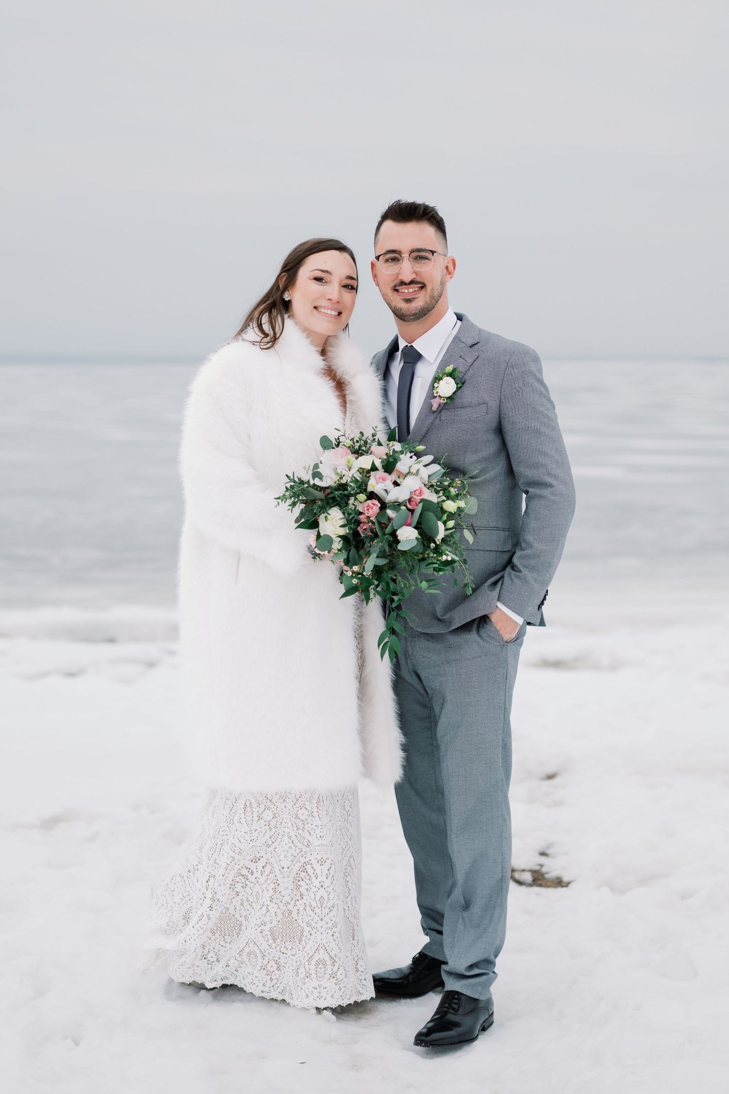 A couple in winter wedding attire poses together on a snowy beach, with the bride holding a pink and green bouquet.