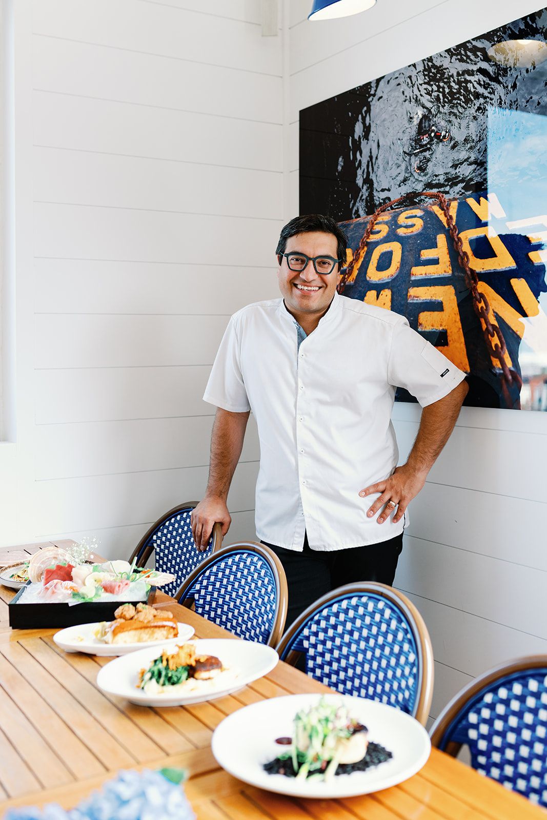 A person standing at a dining table with plated meals and blue bistro chairs in a bright modern setting.