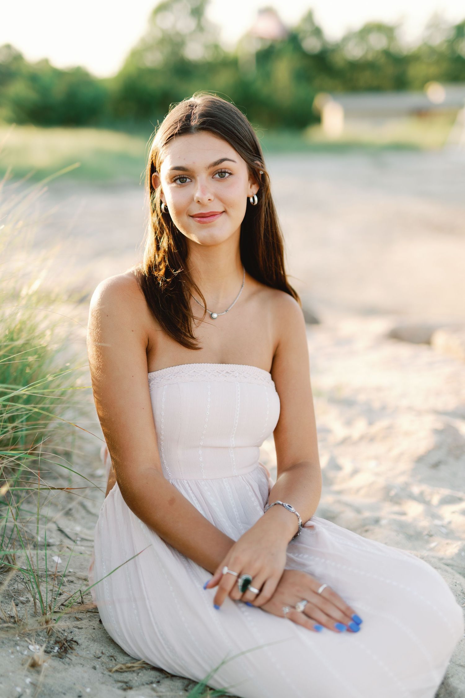 A person in a white strapless dress sits on a sandy beach with tall grass in the background during golden hour.