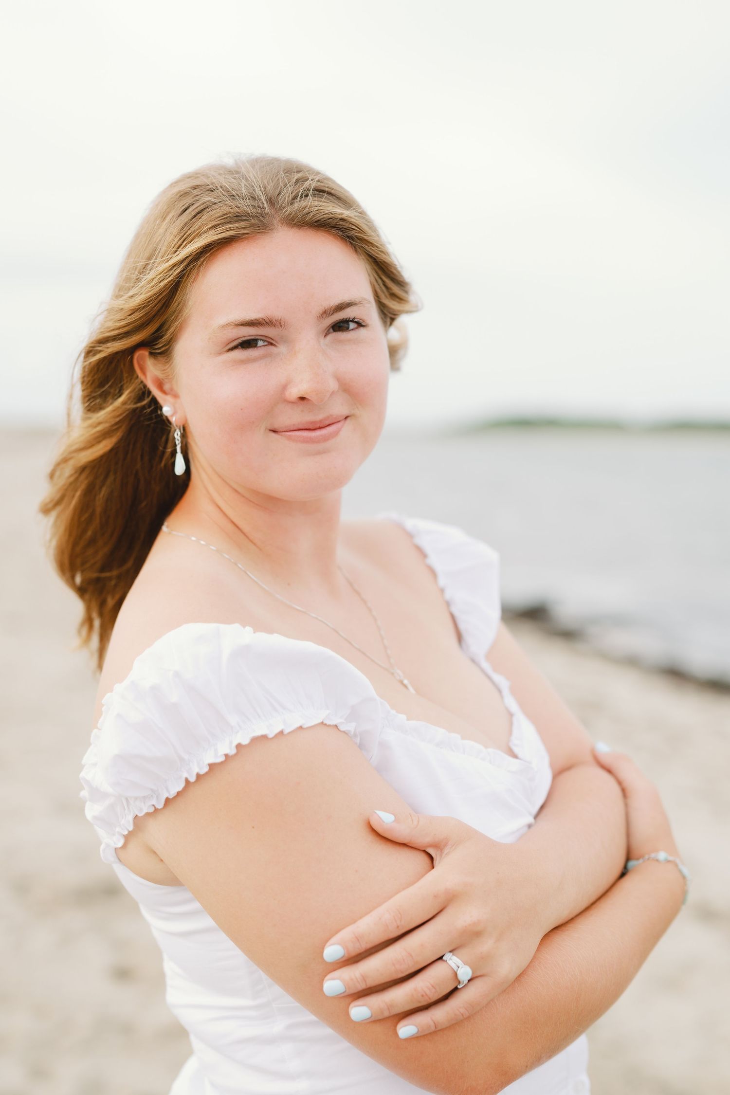 Young woman in white off-shoulder dress smiling at beach, wearing drop earrings and ring.