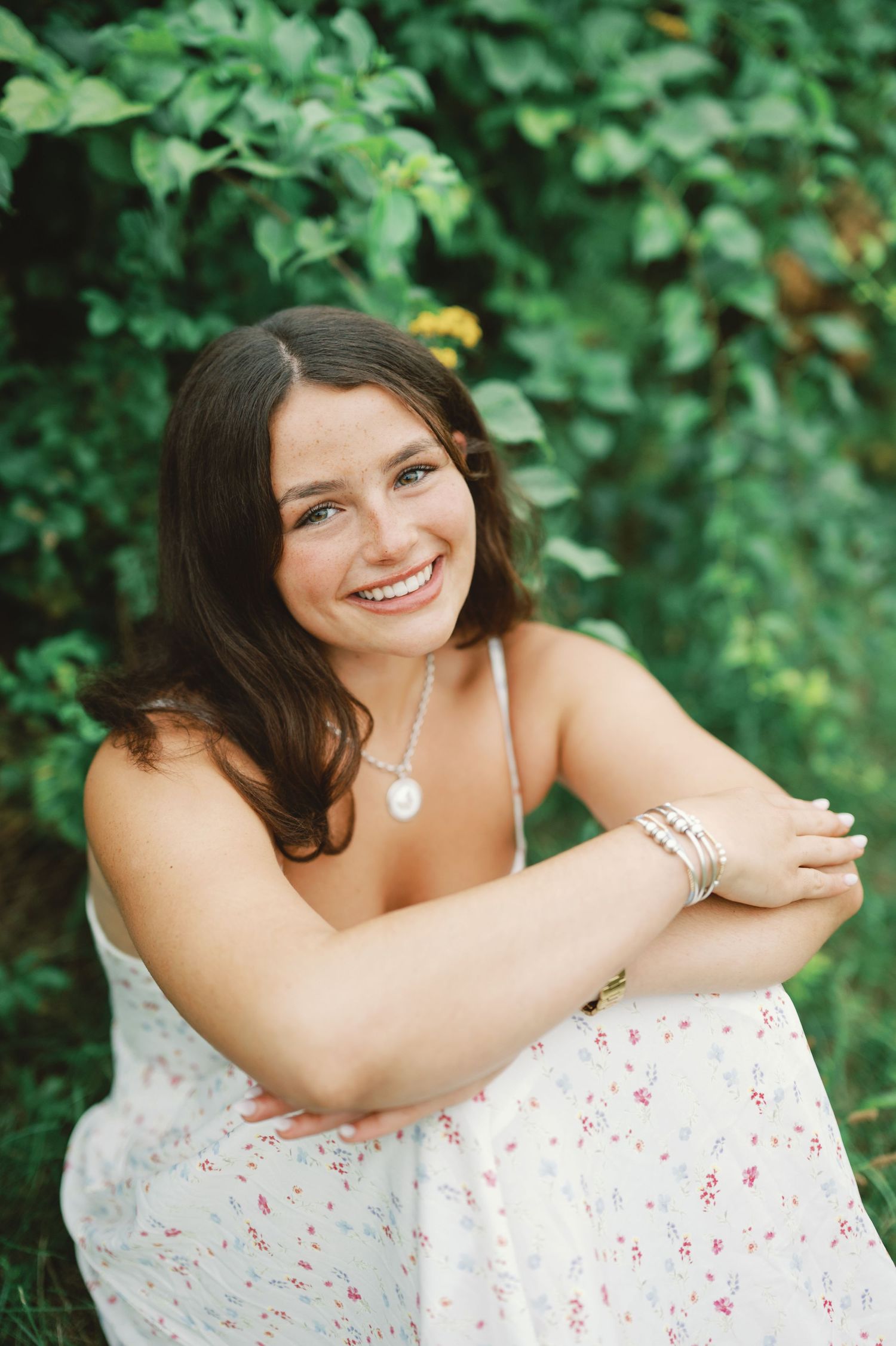 Smiling young woman in floral dress with pearl necklace sitting outdoors against lush green foliage backdrop.