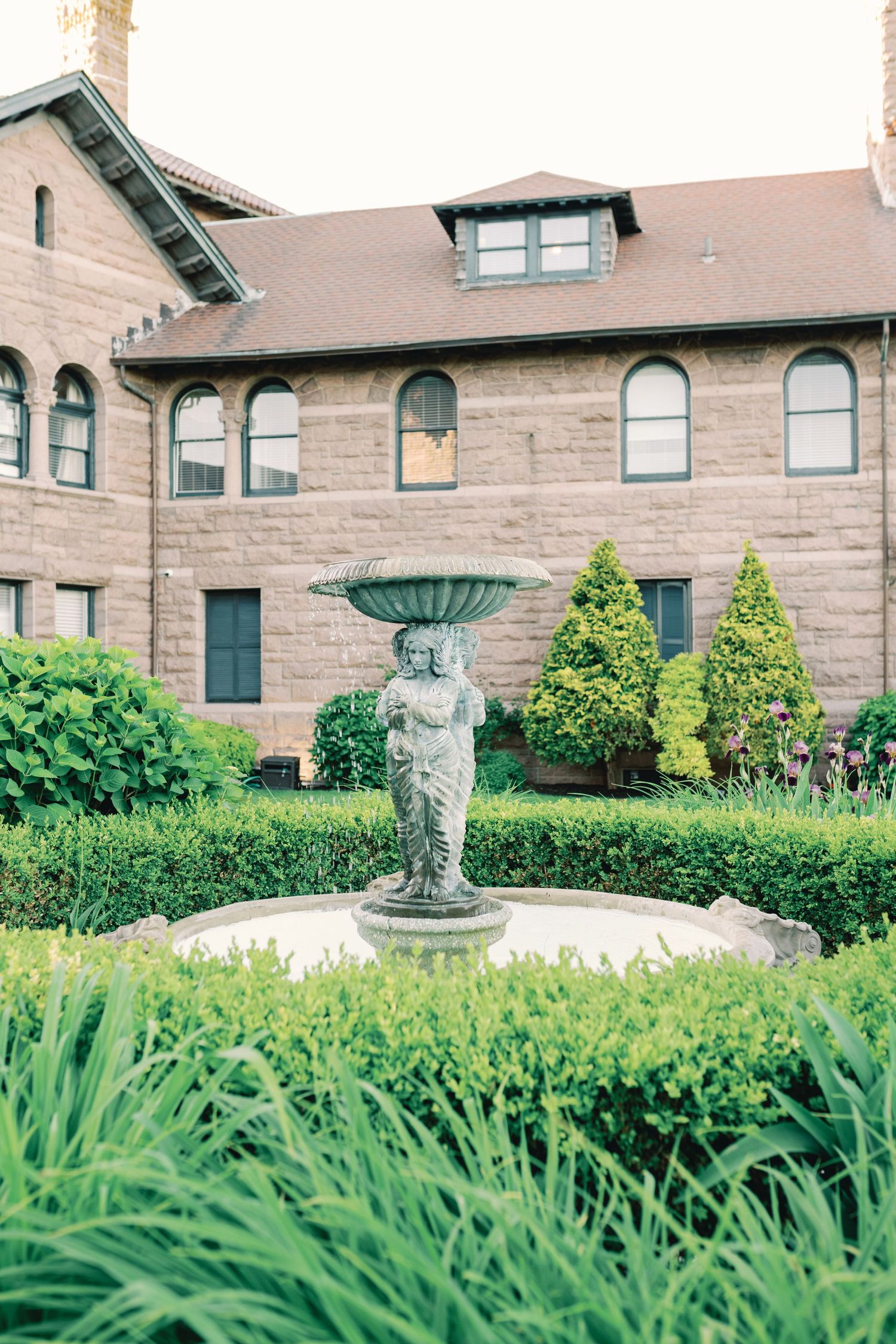 Stone cherub fountain adorns a lush garden with ornate architecture and green landscaping in the background.
