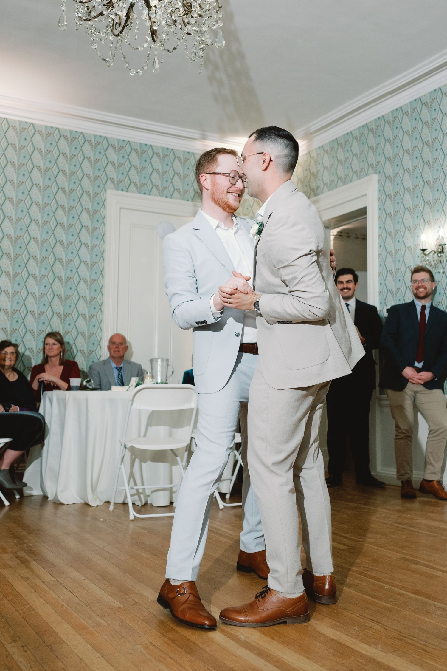 Two people in light colored suits share a romantic first dance moment on a hardwood floor at their wedding reception.