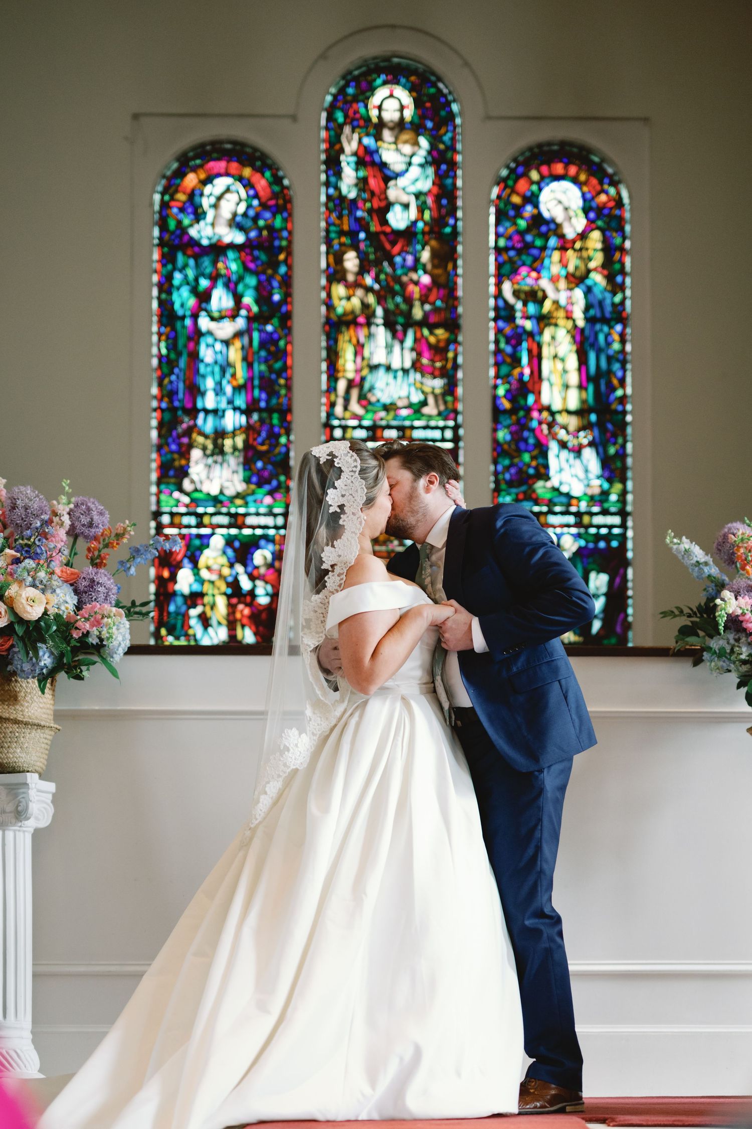 A romantic wedding scene showing a bride and groom embracing in front of stained glass church windows.