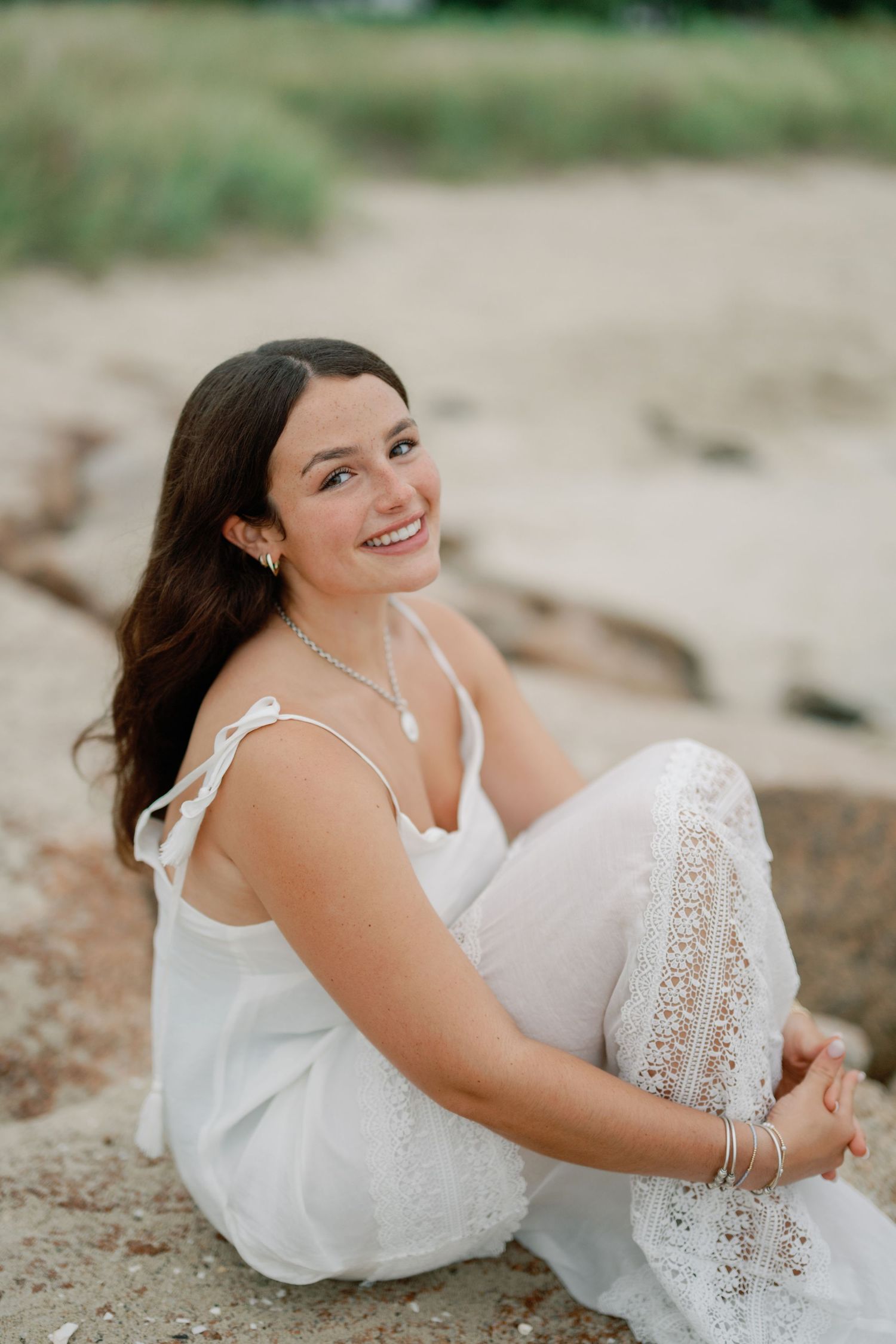 Young woman in white lace dress sitting on sandy beach rocks, smiling joyfully, wearing silver jewelry.